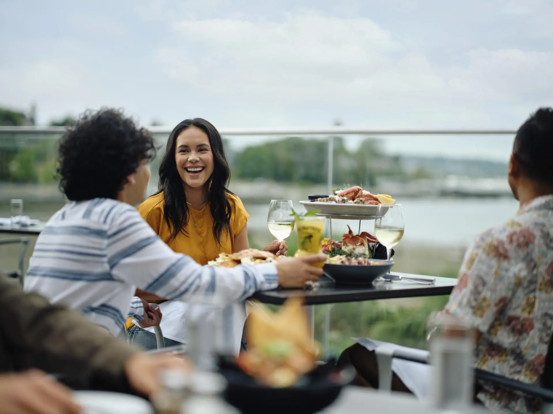 Three people sitting at an outdoor table with seafood dishes and drinks, one woman in a yellow top smiling.