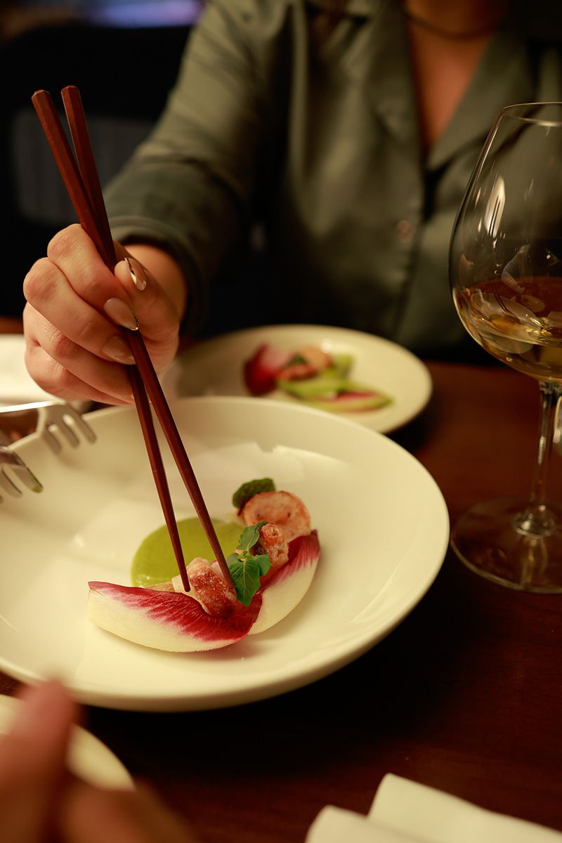 Hand holding chopsticks picking up food from a white plate with a green sauce and red leaf garnish, next to a wine glass.
