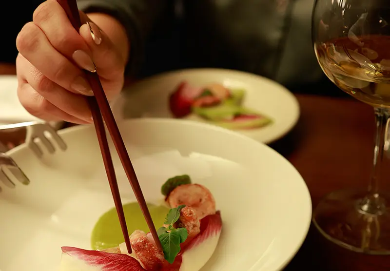 Hand holding chopsticks picking up food from a white plate with a green sauce and red leaf garnish, next to a wine glass.