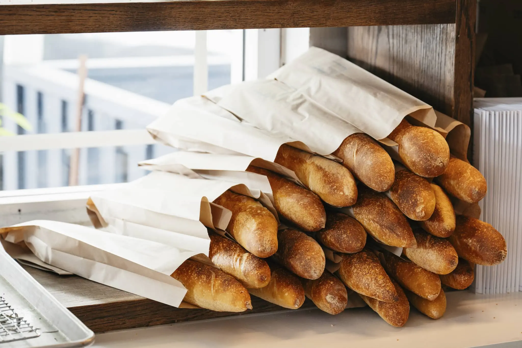 Stack of baguettes partially wrapped in white paper on a wooden shelf near a window.