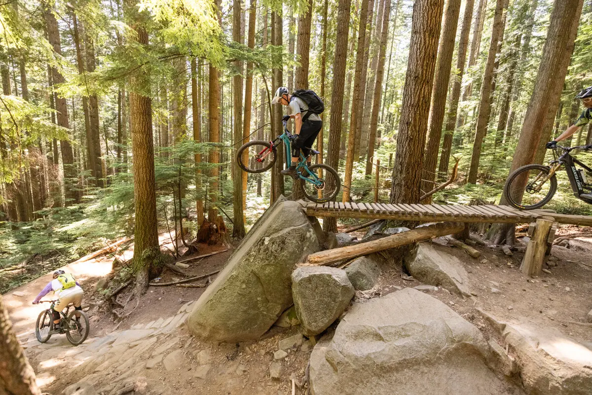 Mountain bikers drop off a steep wood and rock feature in the forest in a still from the film Maneuver Vancouver screening at the Vancouver International Mountain Film Festival