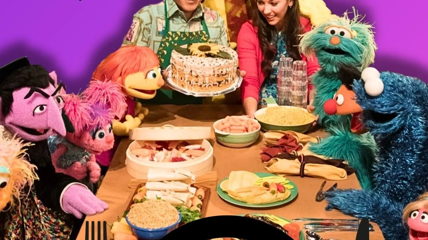 A group of puppets and two people around a table with food, including a cake, chips, and bowls of snacks.