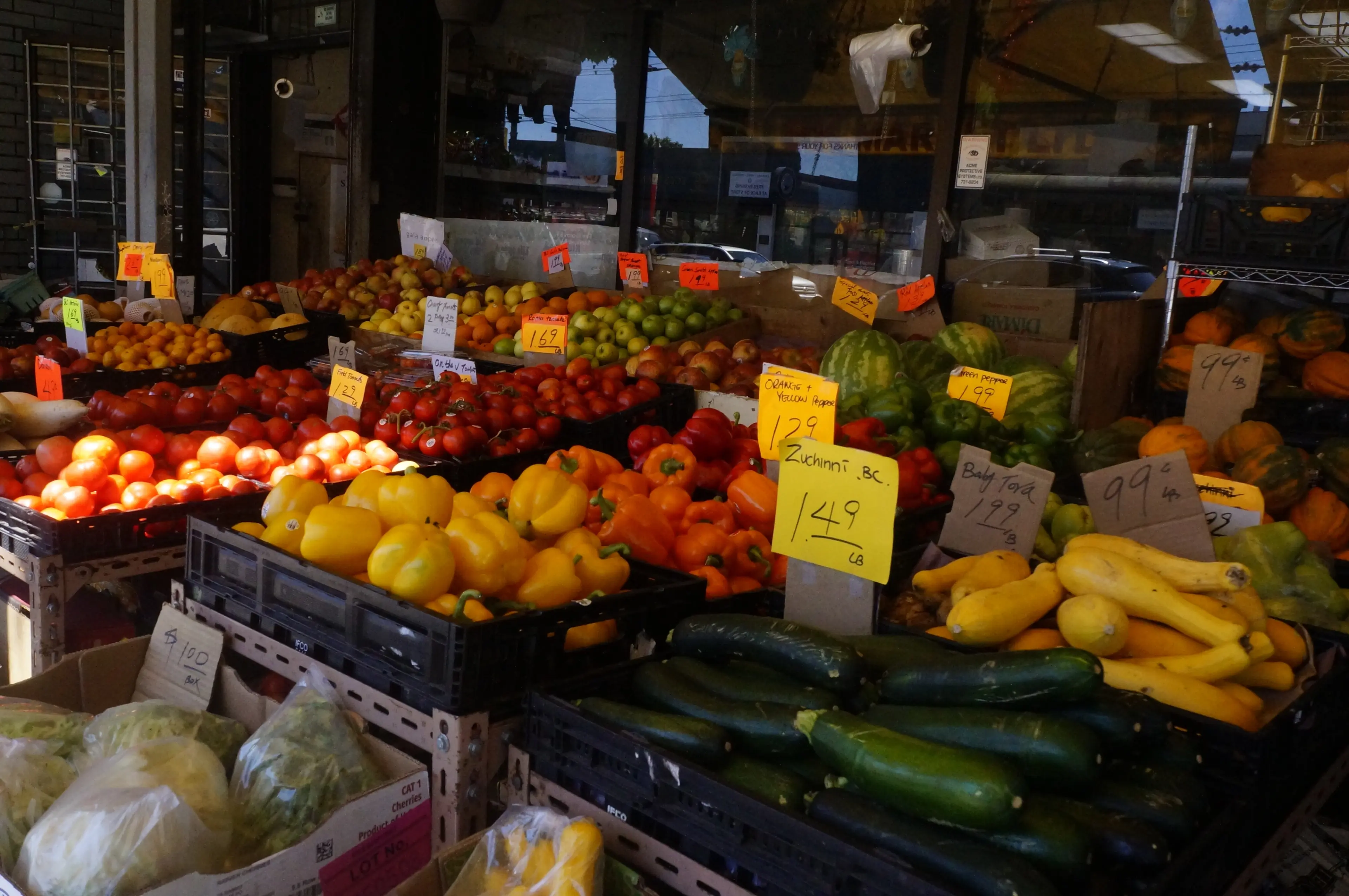 Outdoor produce stand with tomatoes, bell peppers, squash, apples, and handwritten price signs.
