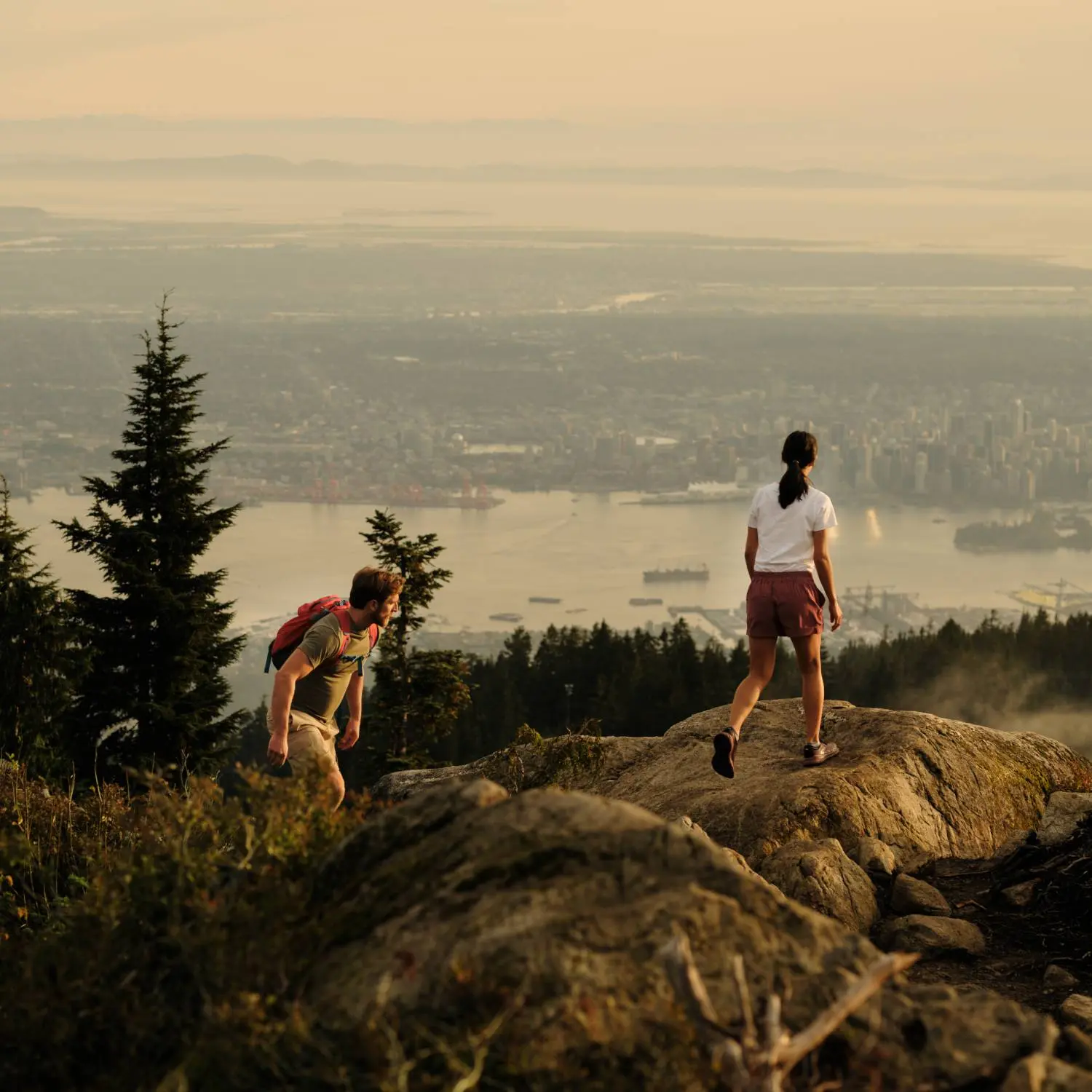 Two hikers on rocky terrain overlooking a city and water at sunset, with trees nearby.