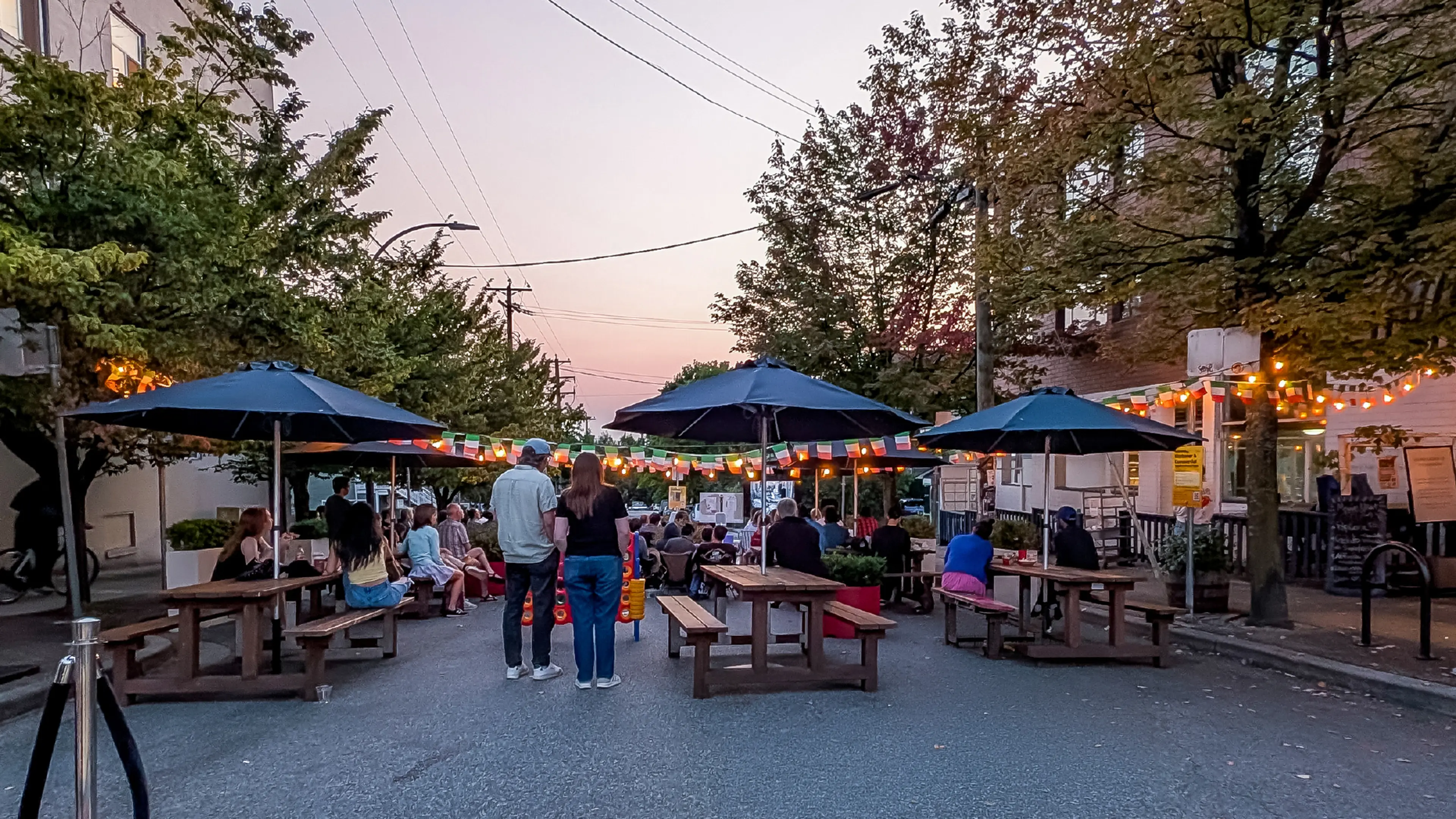 People sitting and standing around picnic tables with umbrellas on a street decorated with string lights and flags at dusk.