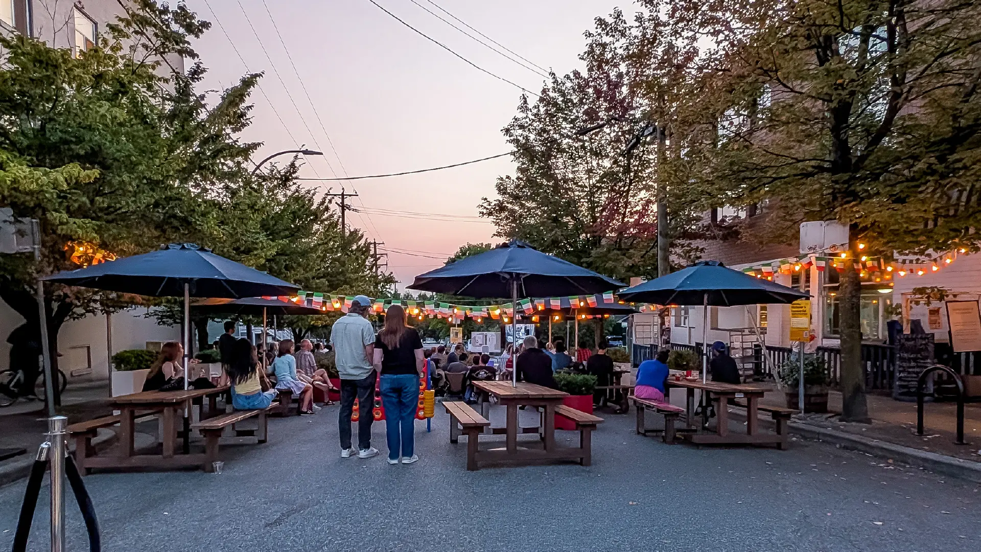 People sitting and standing around picnic tables with umbrellas on a street decorated with string lights and flags at dusk.