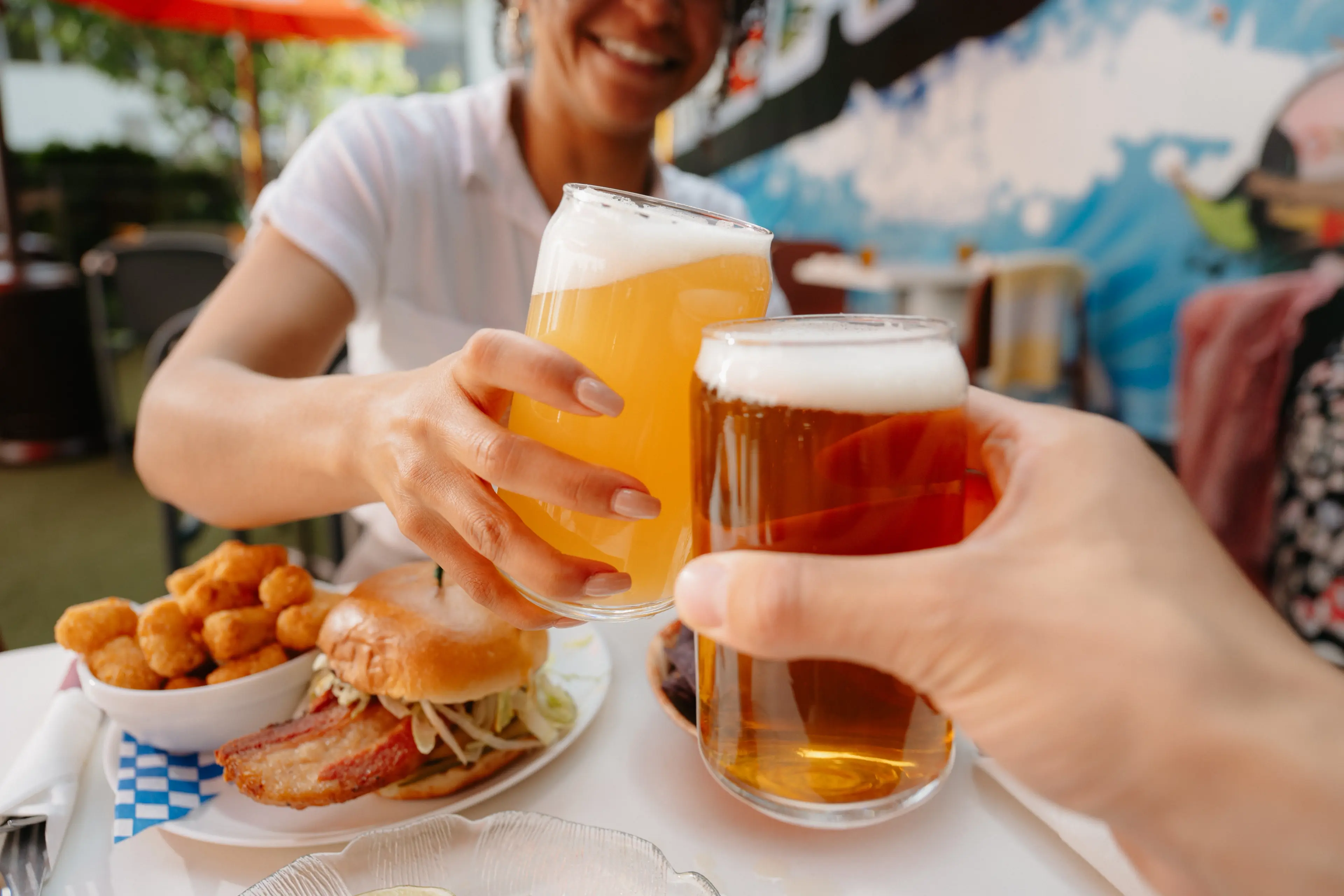 Two people clink beer glasses on a sunny brewery patio, with a plate of burger and tater tots on the table and a colorful mural in the background.