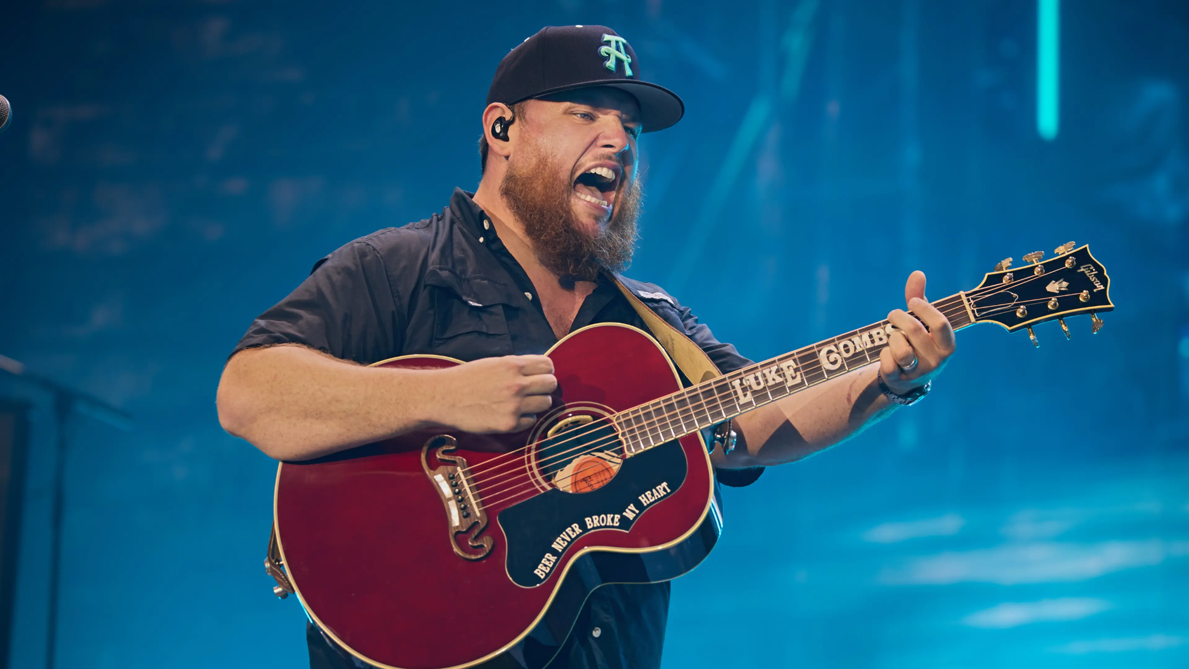 A person wearing a black cap and shirt passionately playing a red acoustic guitar on stage with blue lighting.