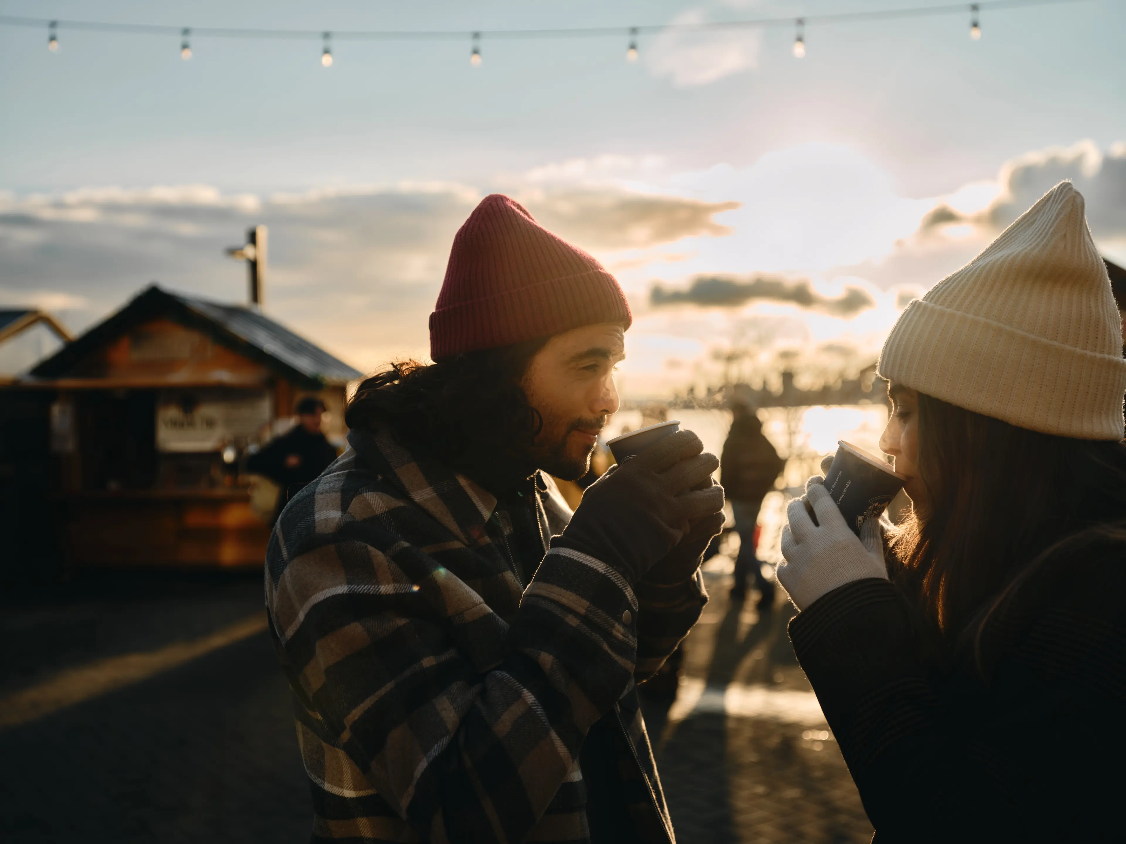 Two people drinking hot beverages in mugs at the North Vancouver Shipyards Christmas Market with the ocean in the background.