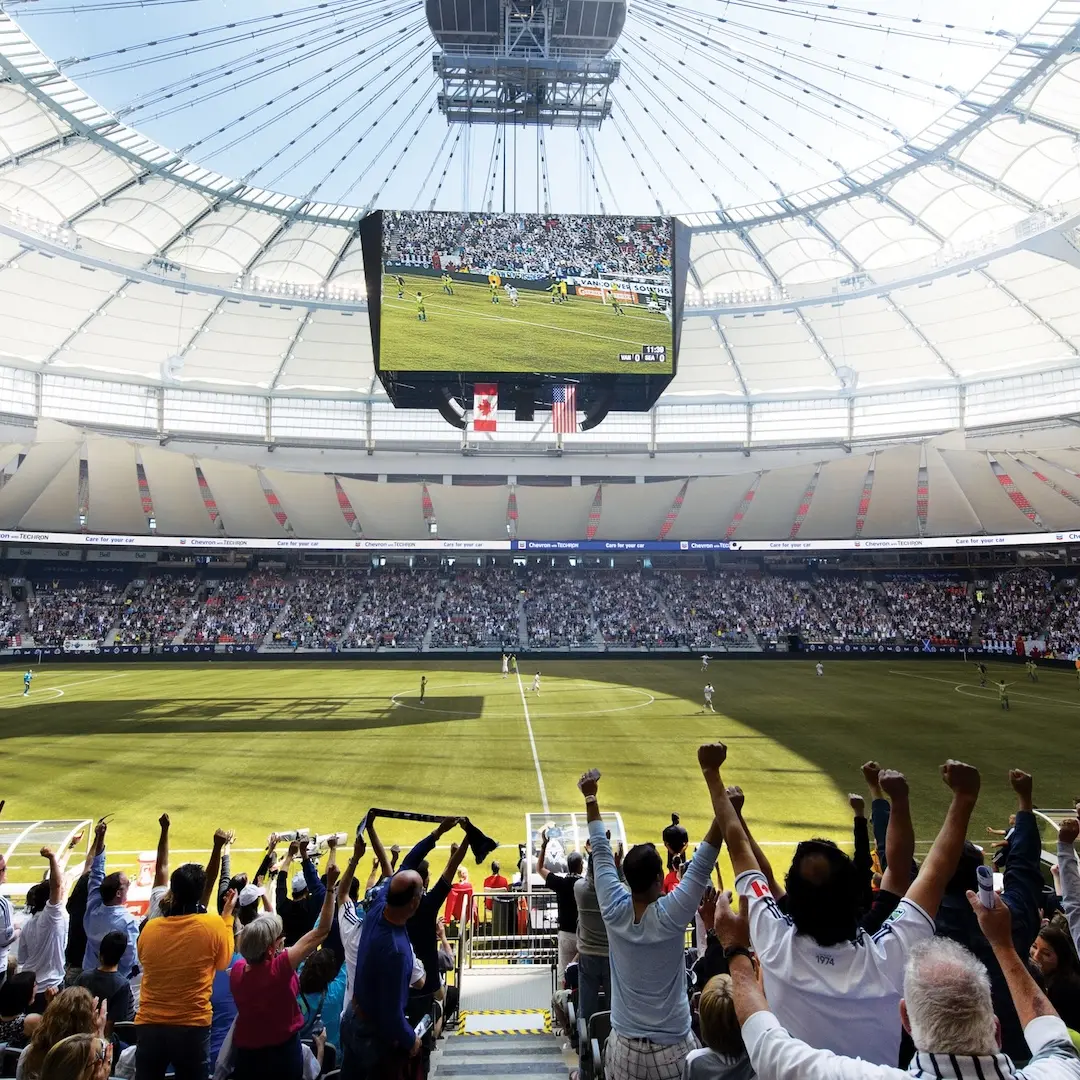 Crowd cheering with raised arms at a soccer stadium during a daytime match under a large white roof.