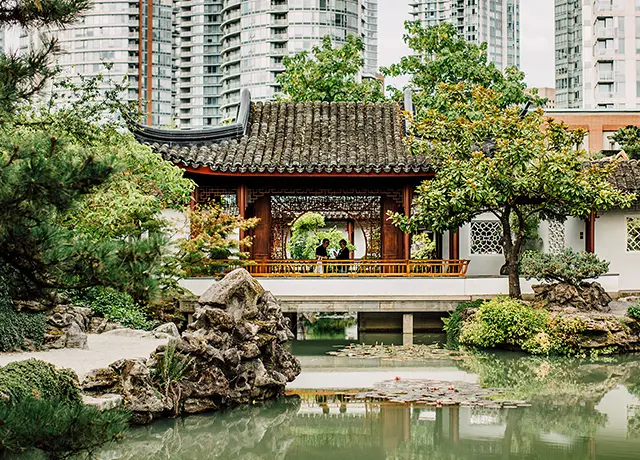 Traditional Chinese garden pavilion with curved roof by a pond, surrounded by trees and rocks, with modern buildings in the background.