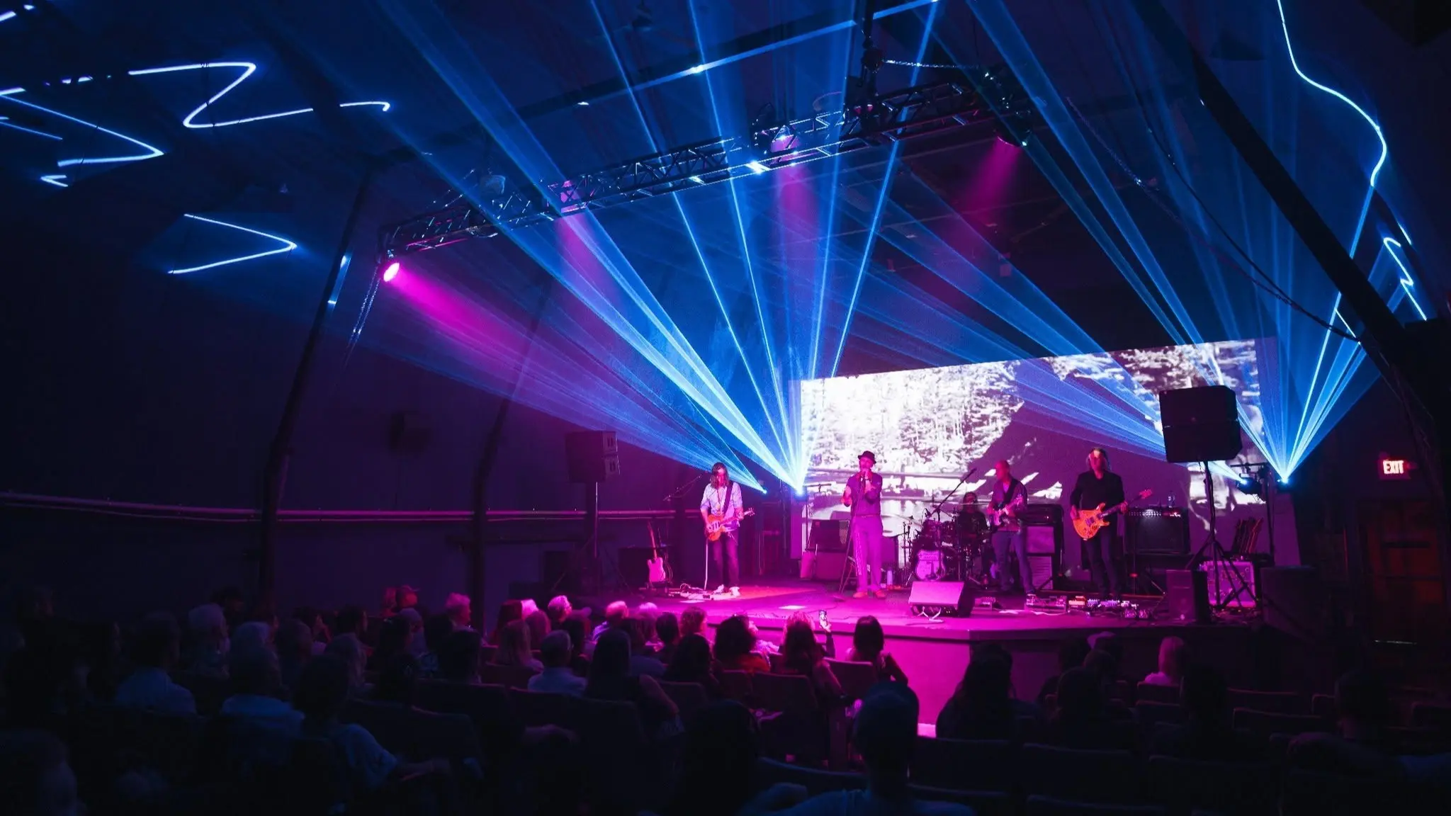 Band performing on stage with blue and purple lights and a large screen behind them, audience seated in front.