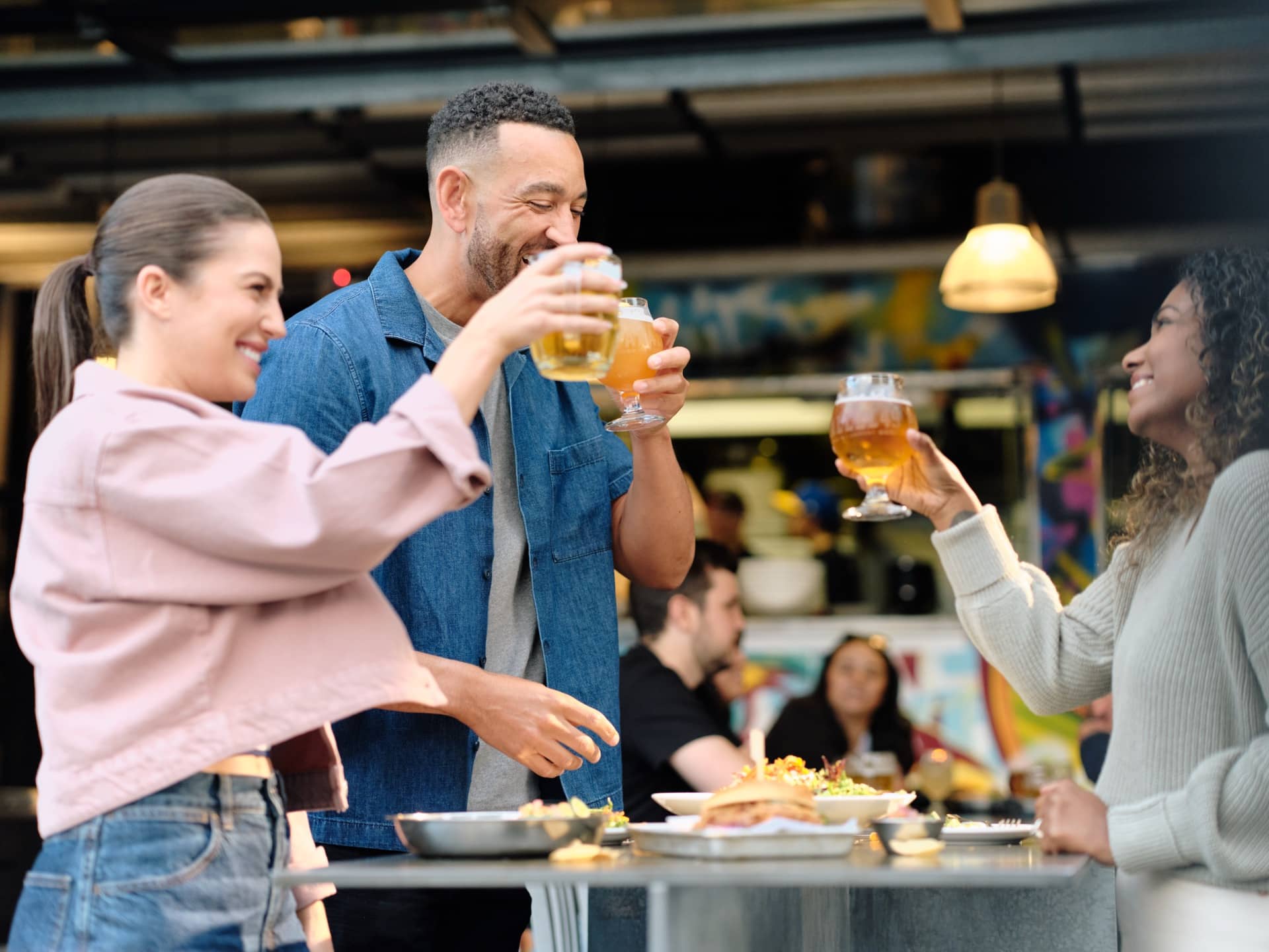 Three people smiling and toasting with glasses of beer around a table with food in a lively indoor setting.