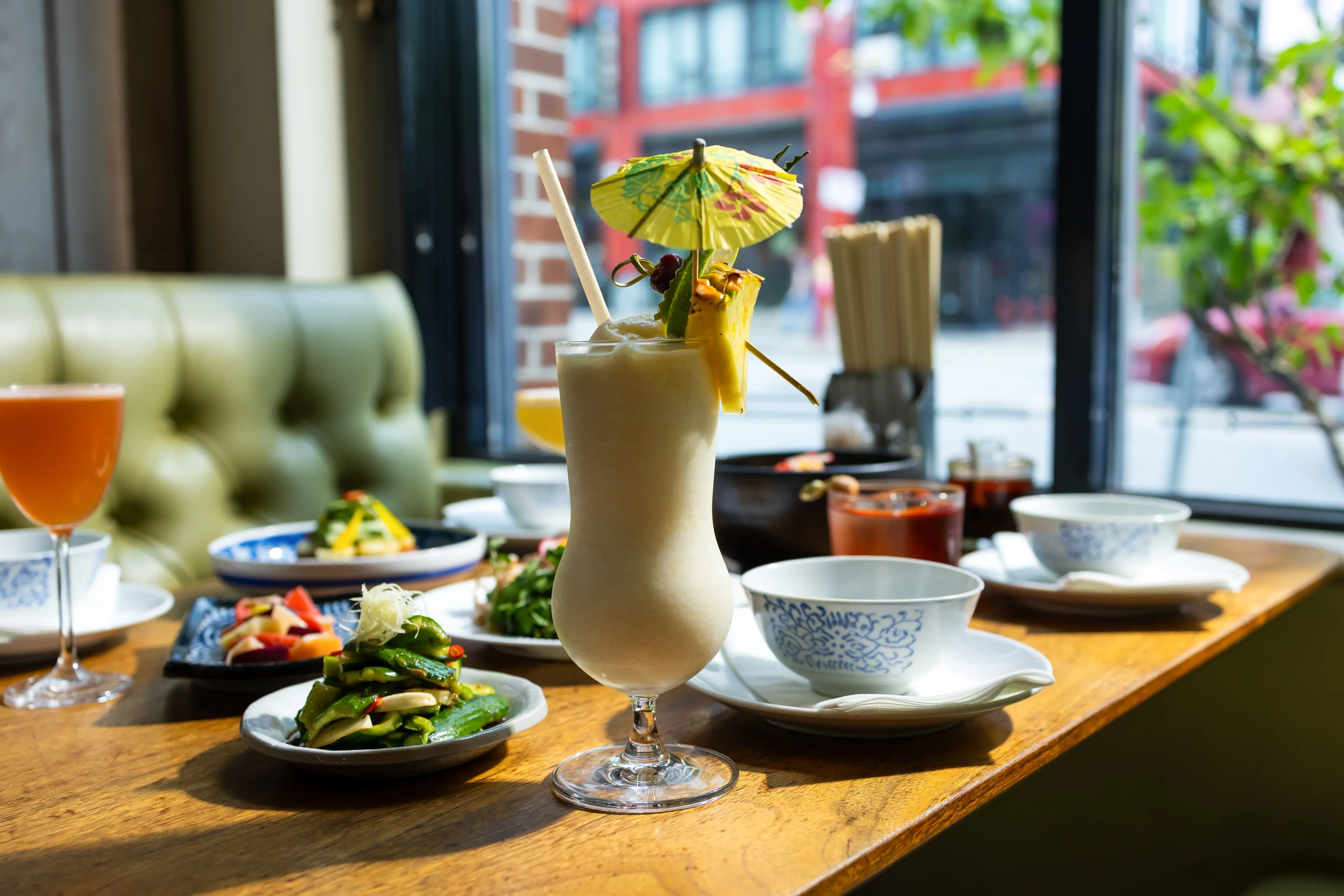A creamy cocktail with a paper umbrella and pineapple garnish on a wooden table with various dishes and drinks near a window.