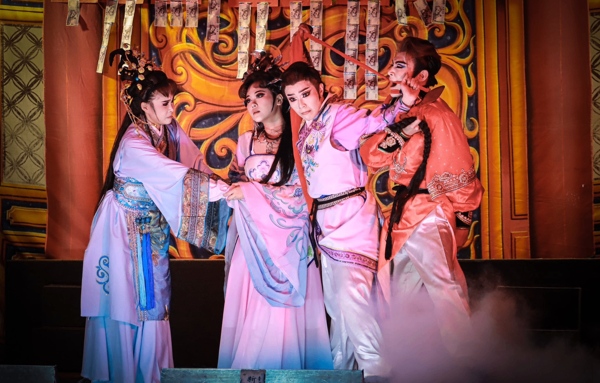 Four people in traditional Asian costumes performing a dramatic scene on stage with ornate backdrop and smoke effects.