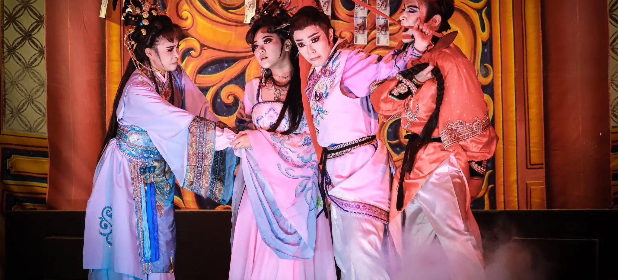 Four people in traditional Asian costumes performing a dramatic scene on stage with ornate backdrop and smoke effects.