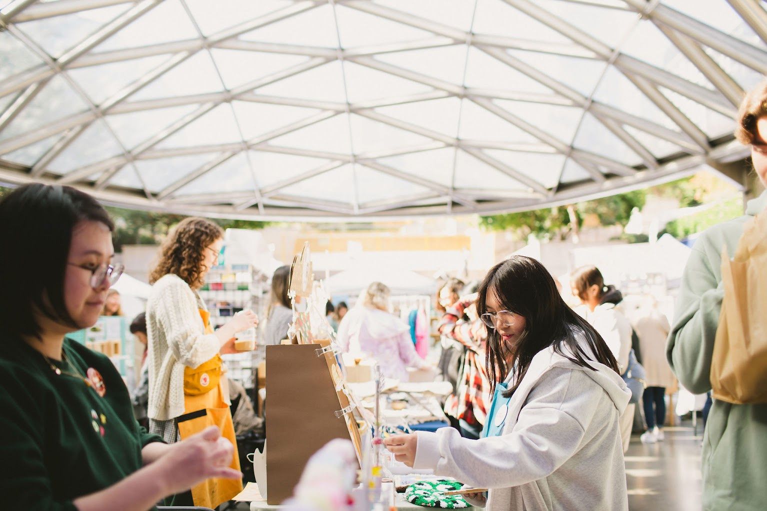 People browsing and shopping at a market stall under a glass roof structure on a sunny day.