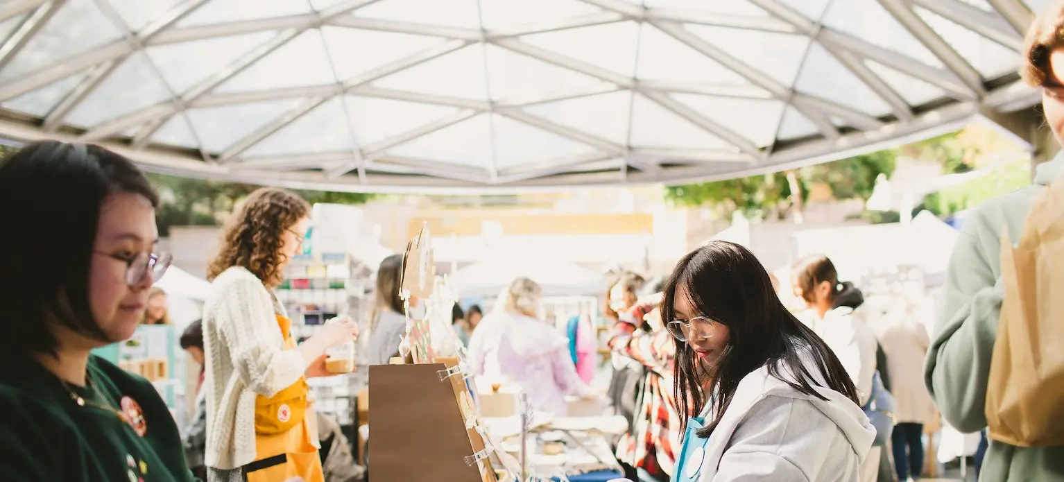 People browsing and shopping at a market stall under a glass roof structure on a sunny day.