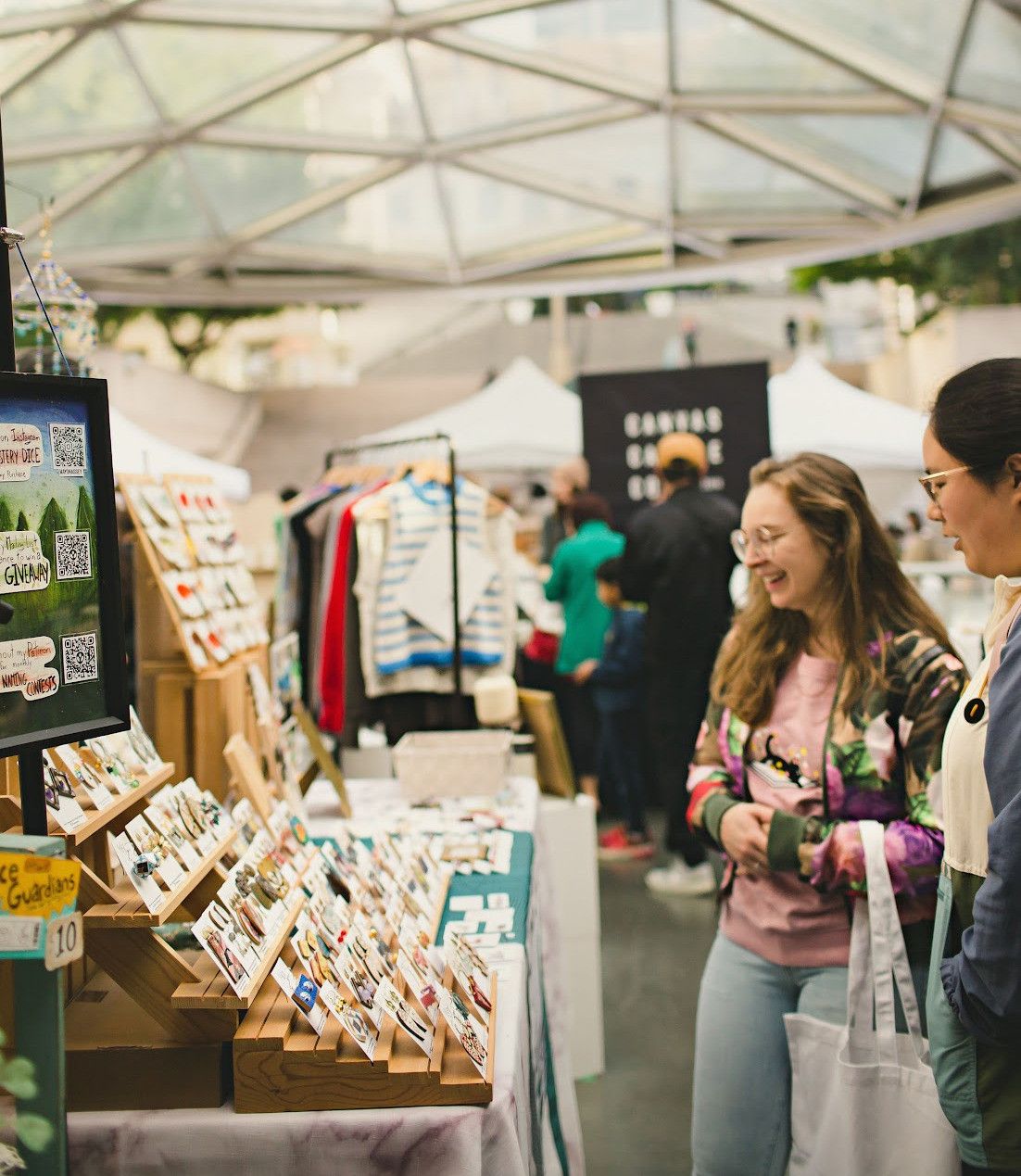 Two people browsing jewelry displayed on wooden stands at an outdoor market under a canopy.