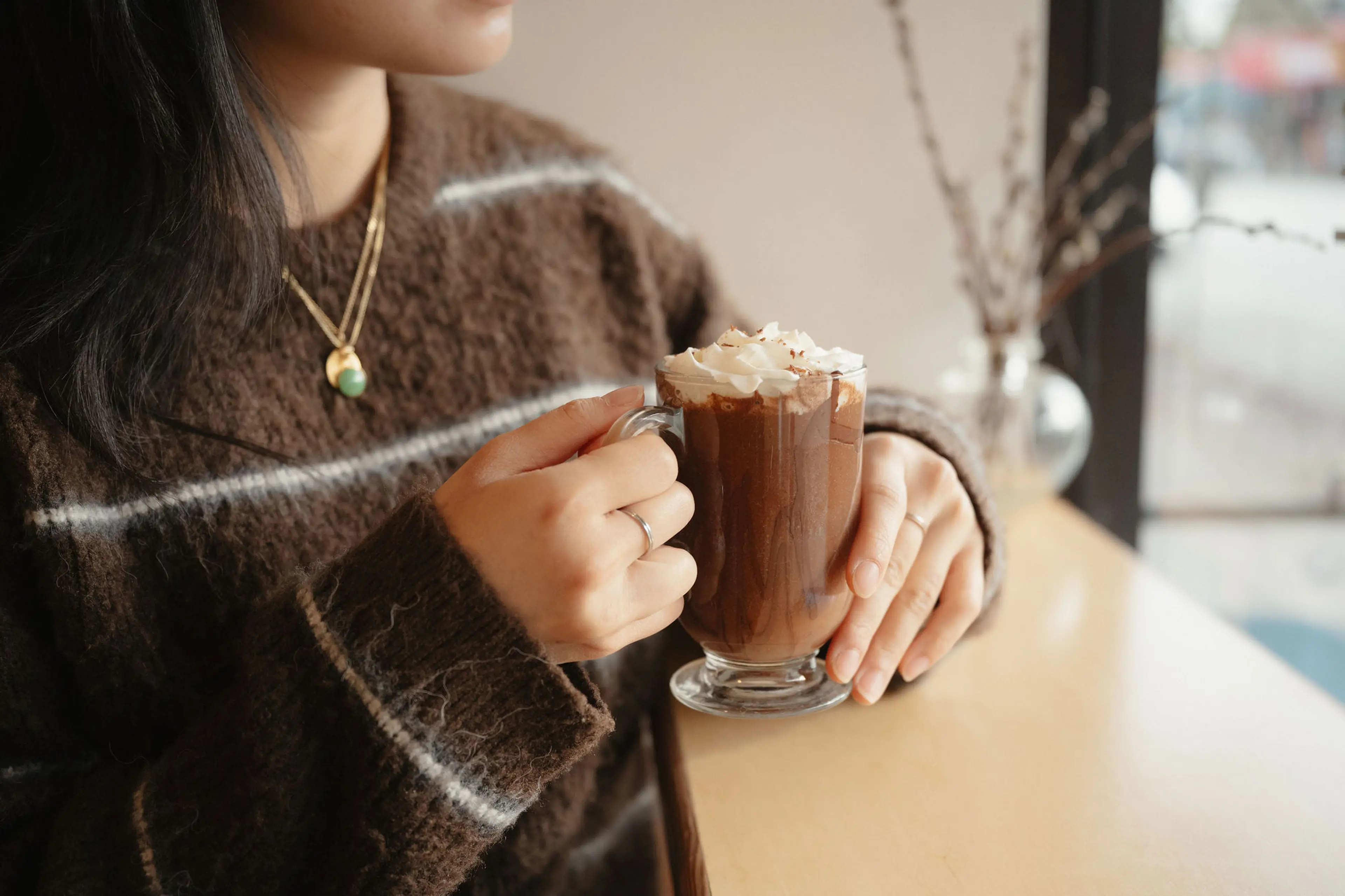 Person in brown sweater holding a glass mug of hot chocolate topped with whipped cream at a table near a window.