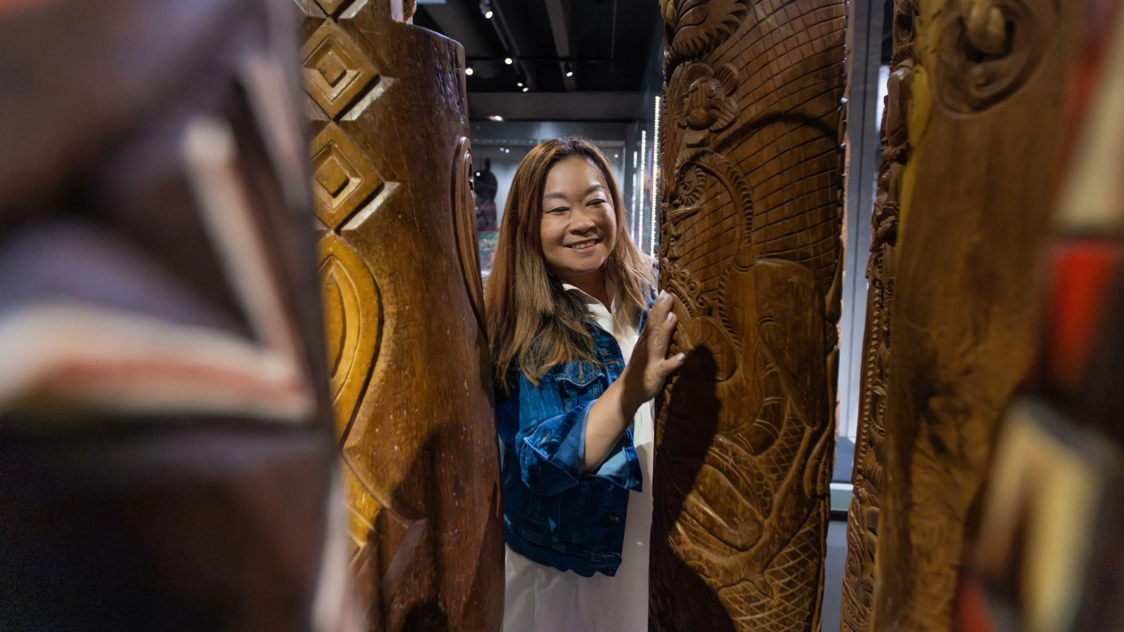 A smiling woman gently touches the textured surface of intricately carved wooden Indigenous poles inside a museum, enjoying a tactile art experience.