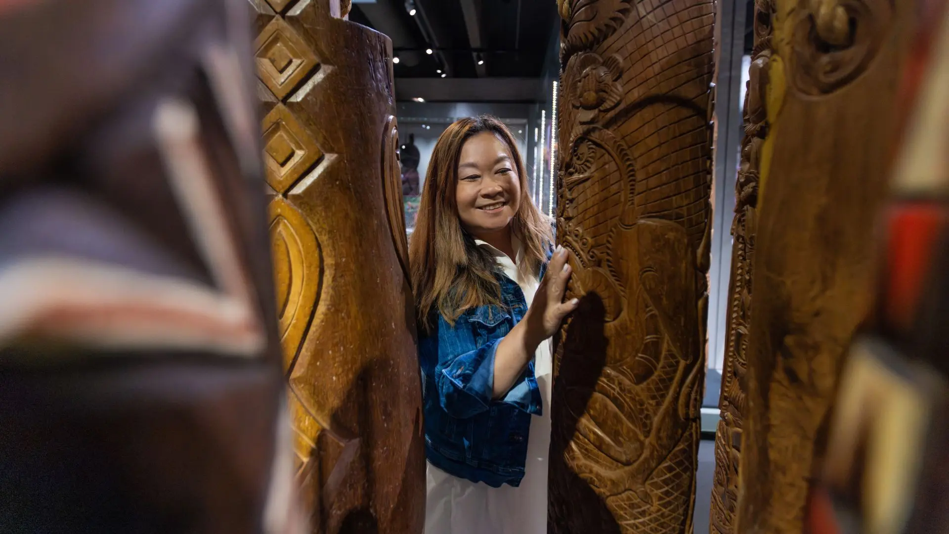 A smiling woman gently touches the textured surface of intricately carved wooden Indigenous poles inside a museum, enjoying a tactile art experience.