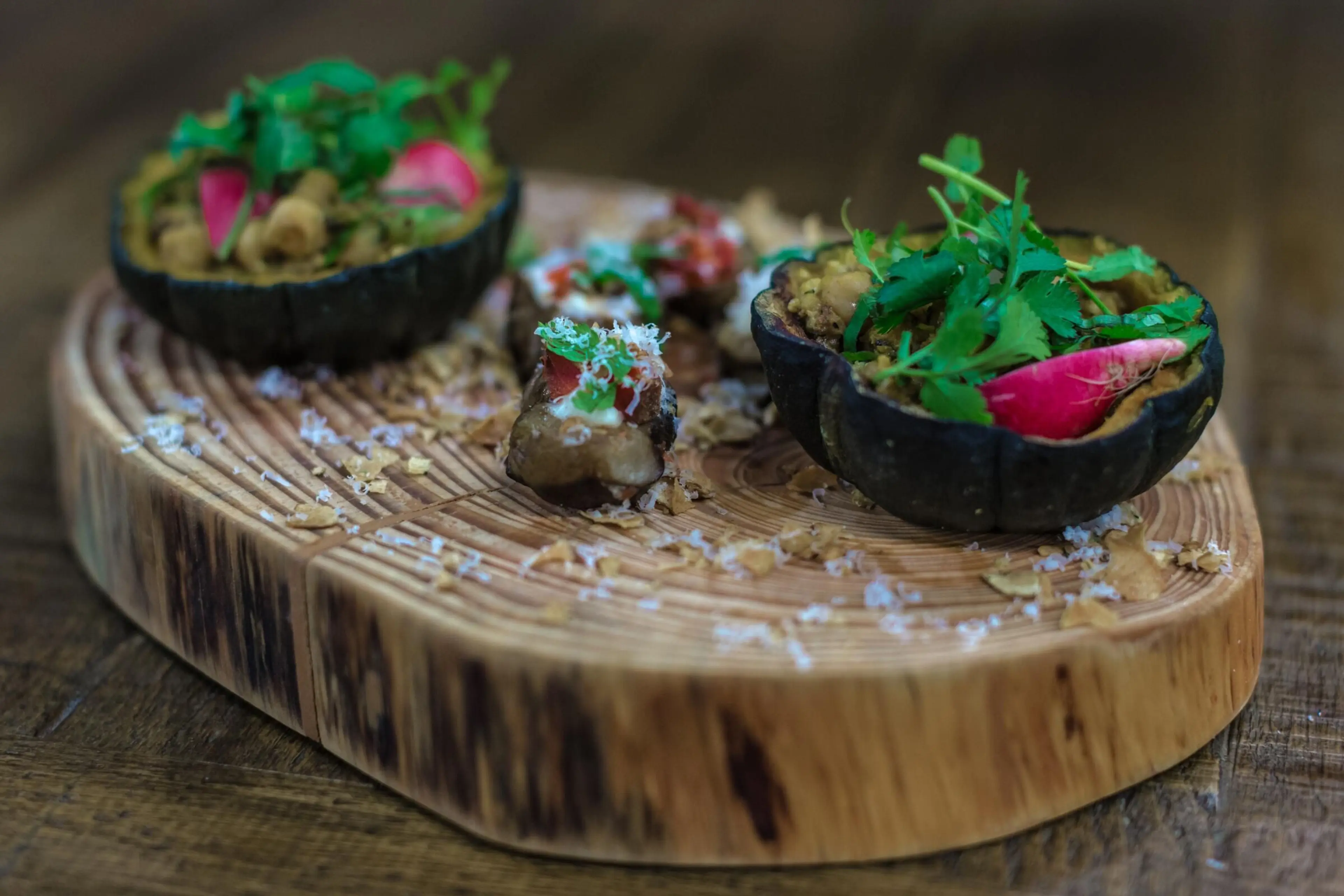 Two black squash halves filled with a creamy dish topped with radish slices and green herbs on a wooden serving board.