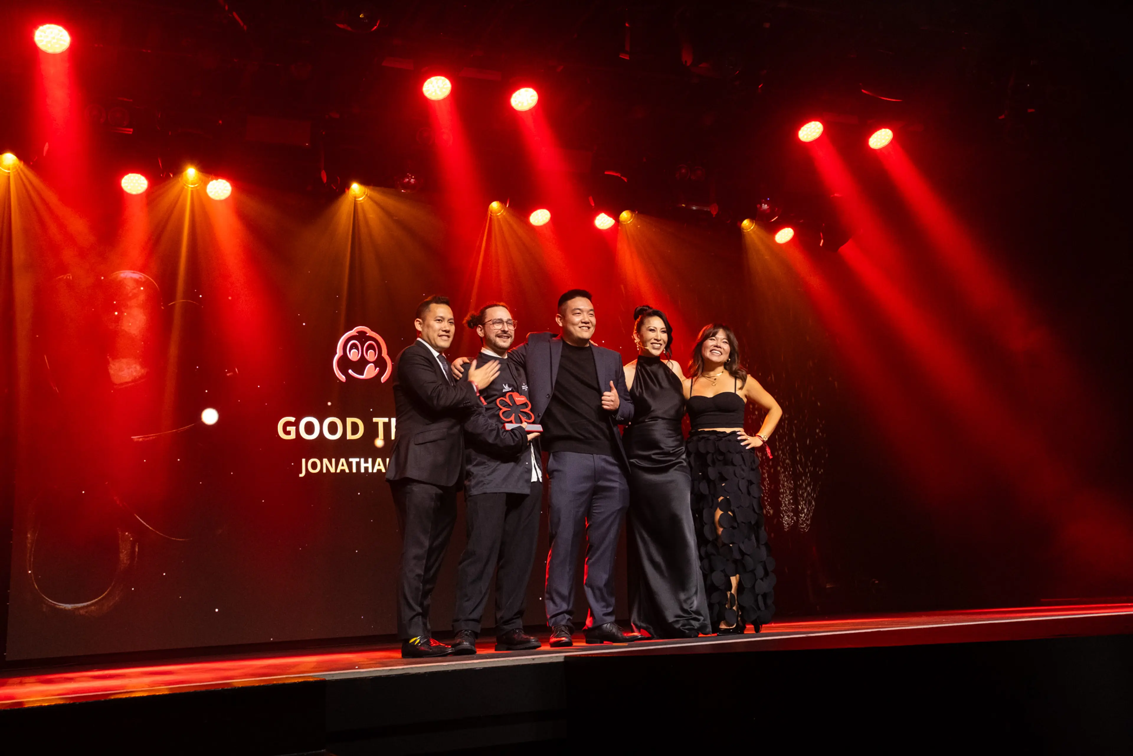 Five people in formal attire standing on stage with red and yellow spotlights, posing for a photo with an award.
