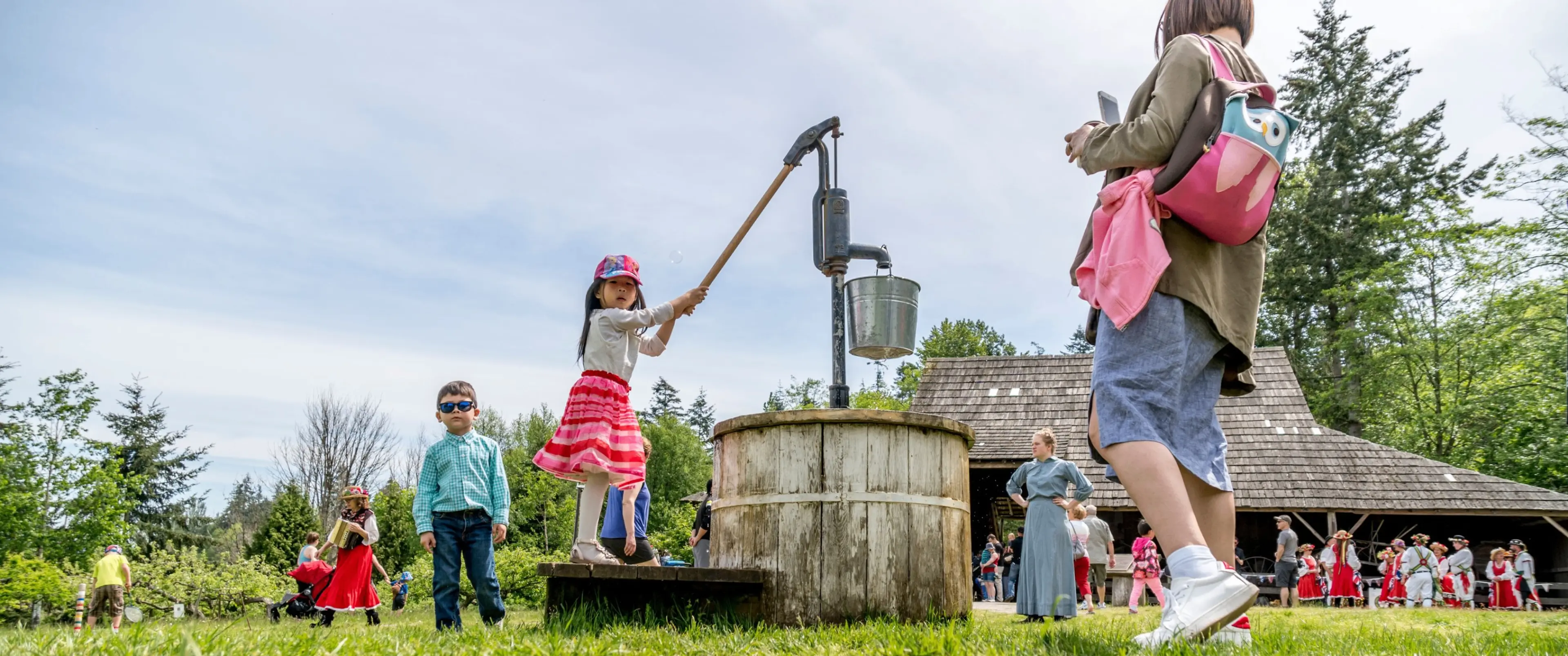 Children and adults outdoors near a wooden water pump and a rustic building, with some children dressed in colorful clothing.
