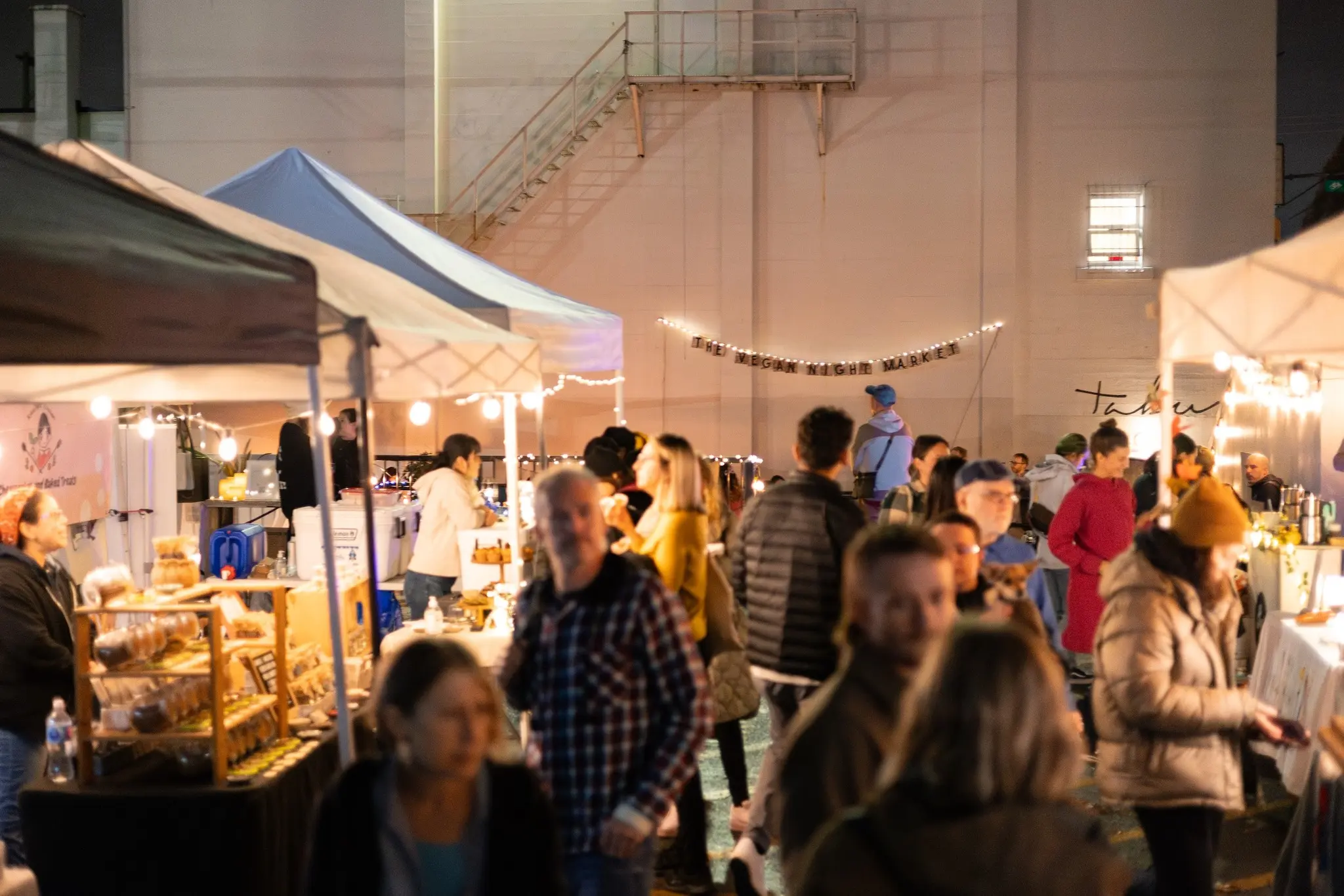 Crowd of people walking and browsing stalls at an outdoor night market with string lights and tents.