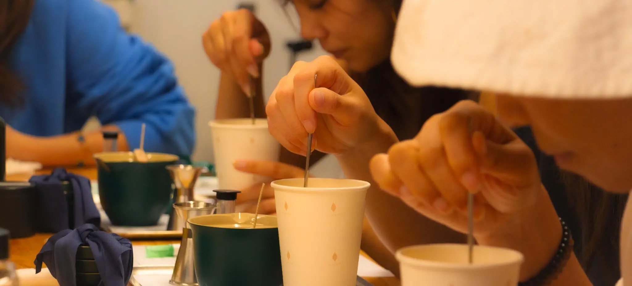Three people stirring drinks in cups on a wooden table with trays and small metal containers.