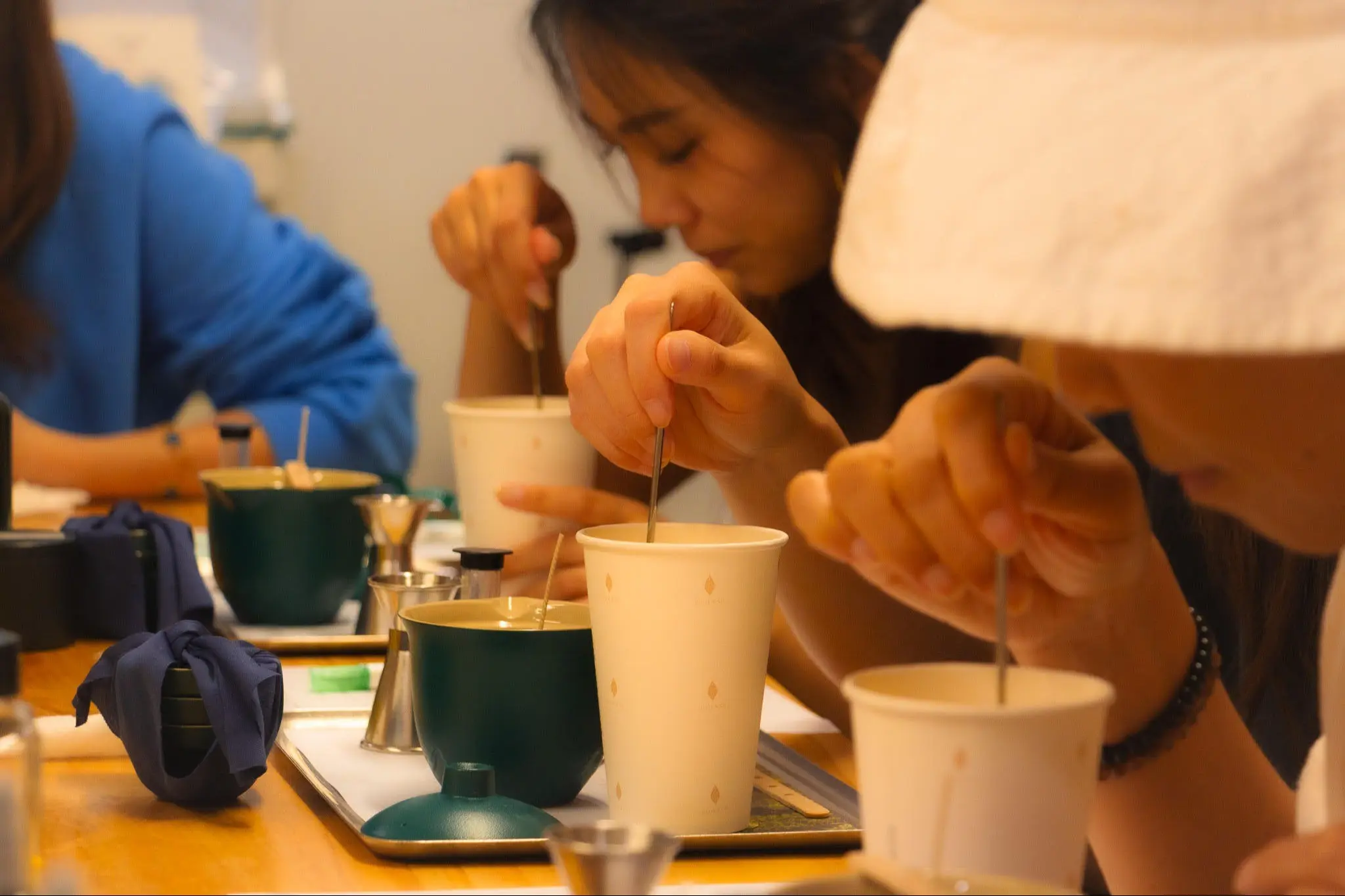 Three people stirring drinks in cups on a wooden table with trays and small metal containers.
