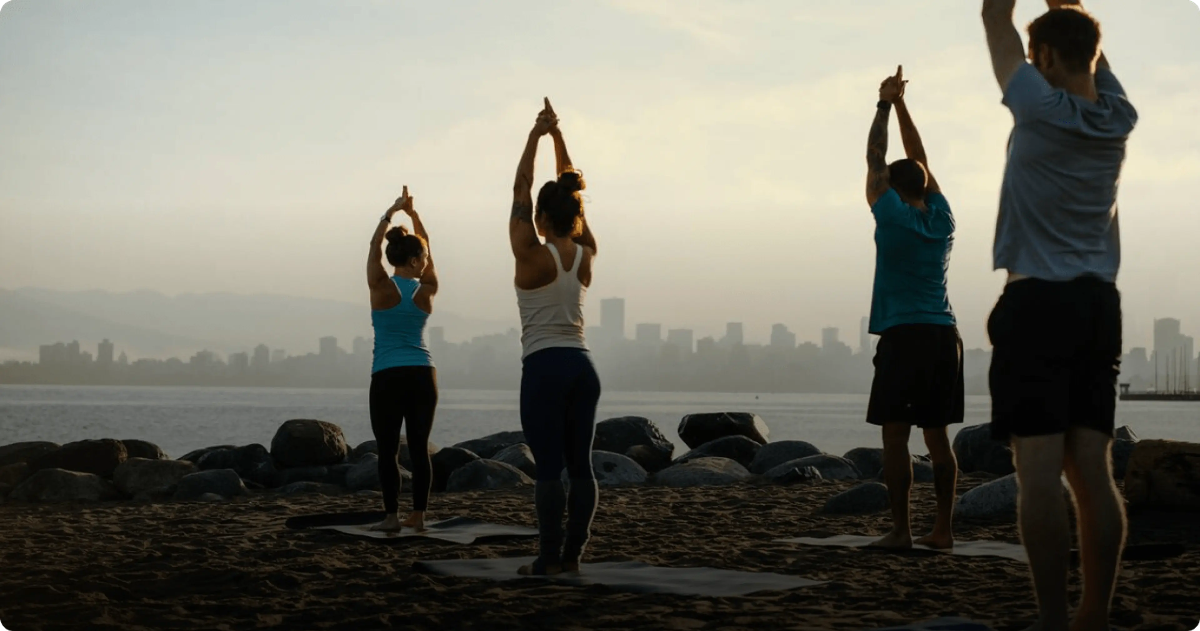A group of people practice yoga on a sandy beach at sunrise—an inspiring start to any Vancouver itinerary—with mats arranged near rocks and the city skyline in the background.