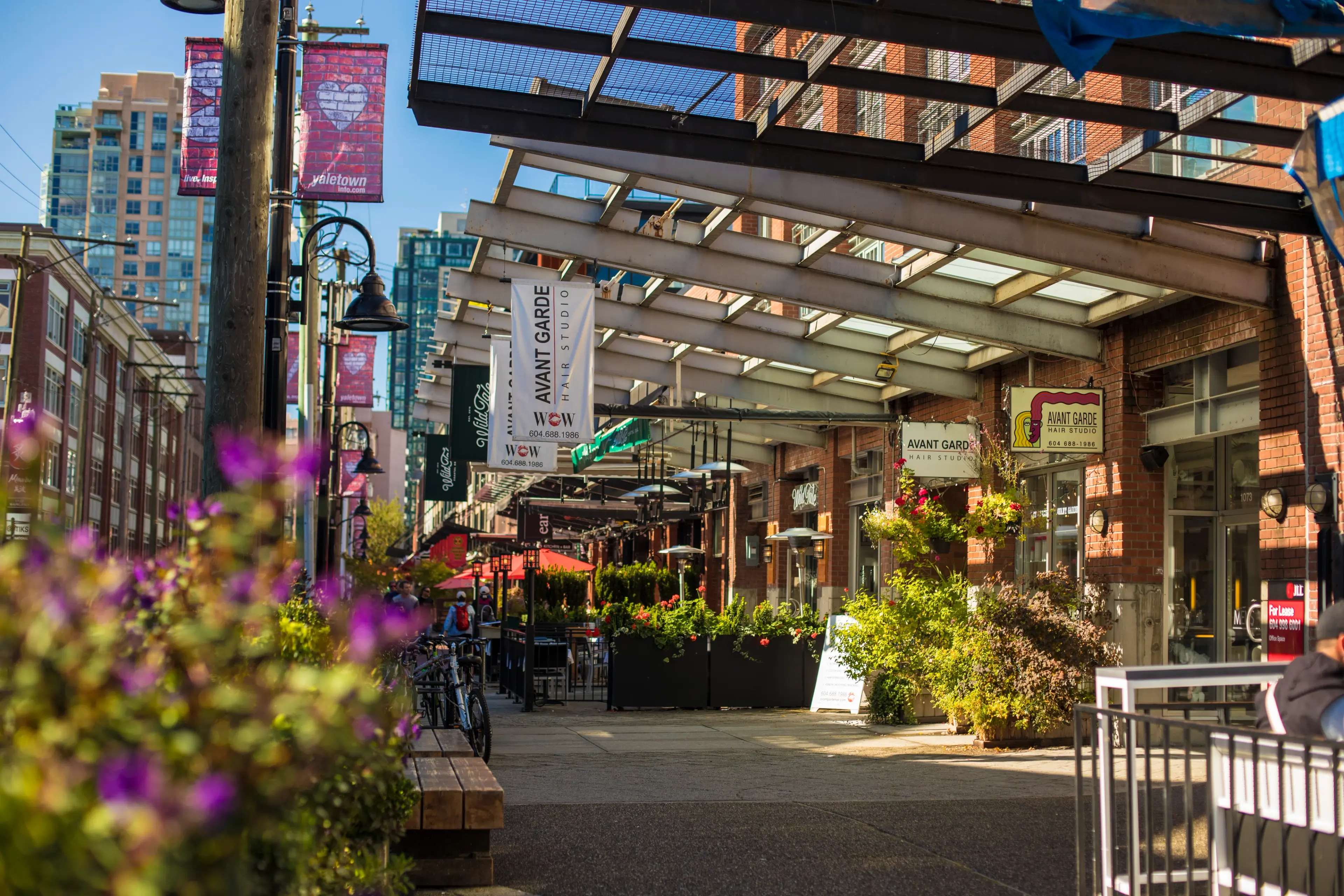 Outdoor urban street scene with brick buildings, hanging signs for Avant Garde Hair Studio, plants, and purple flowers in foreground.