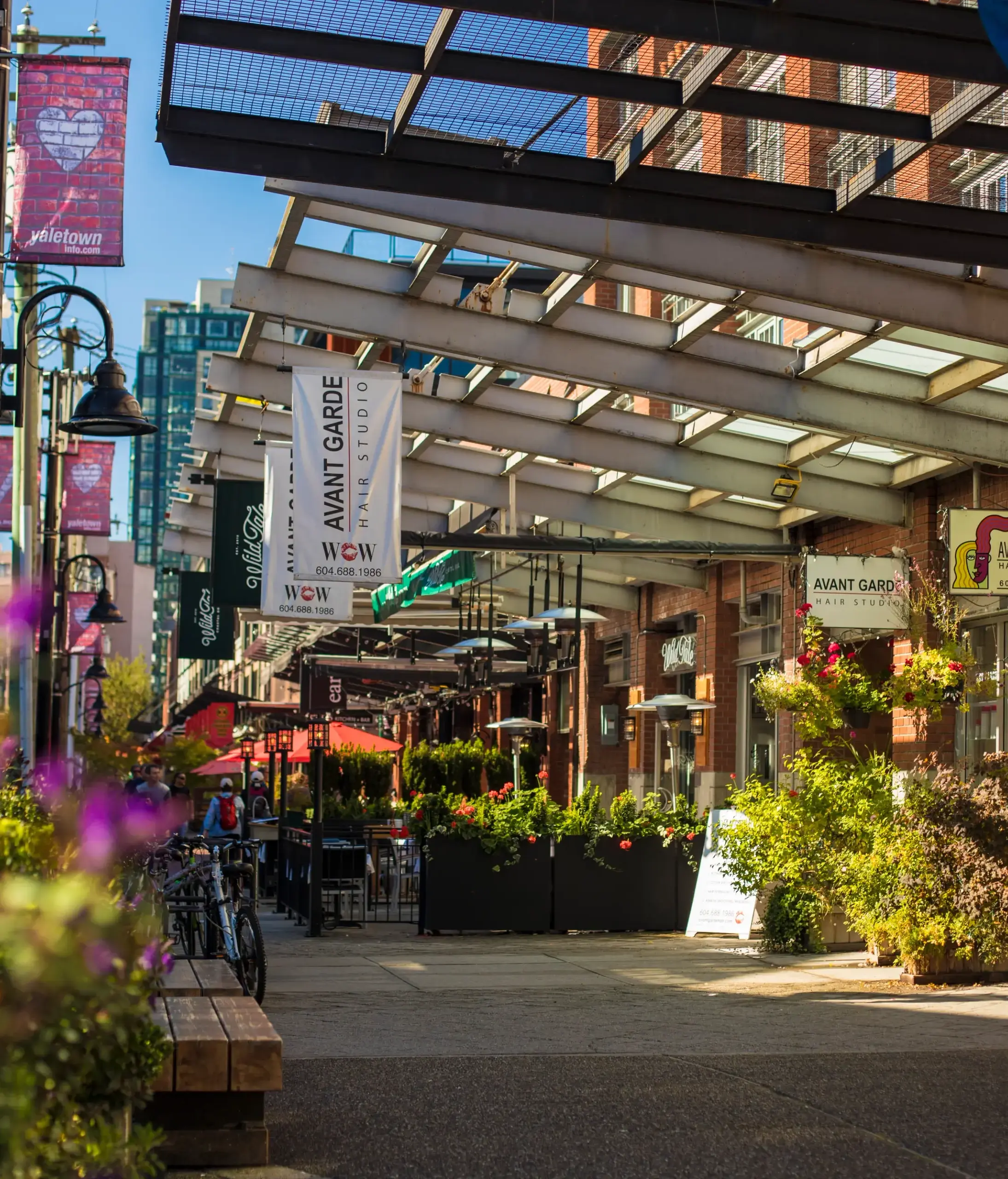 Outdoor urban street scene with brick buildings, hanging signs for Avant Garde Hair Studio, plants, and purple flowers in foreground.