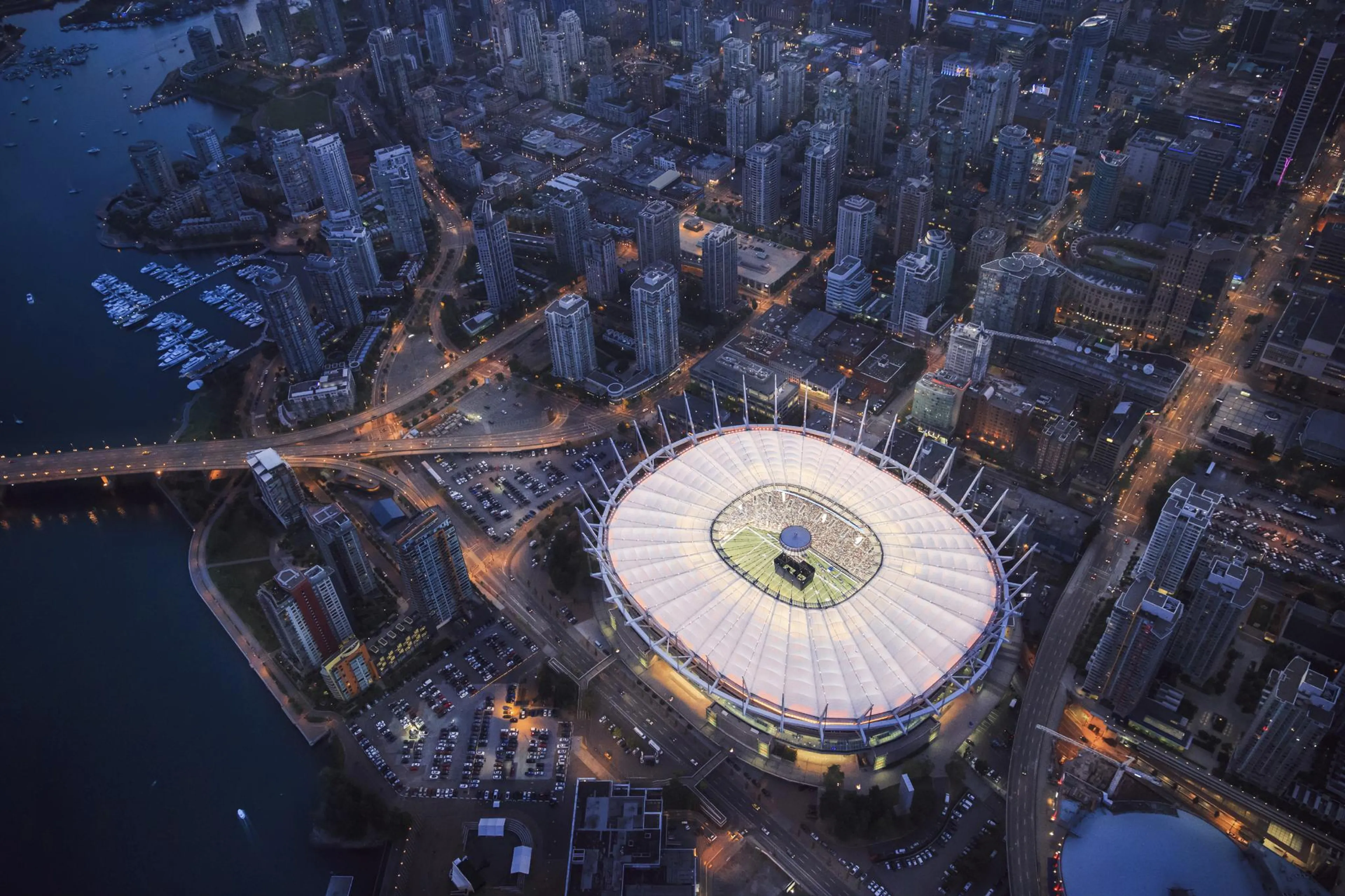 Aerial view of a brightly lit stadium surrounded by city buildings and roads at dusk.