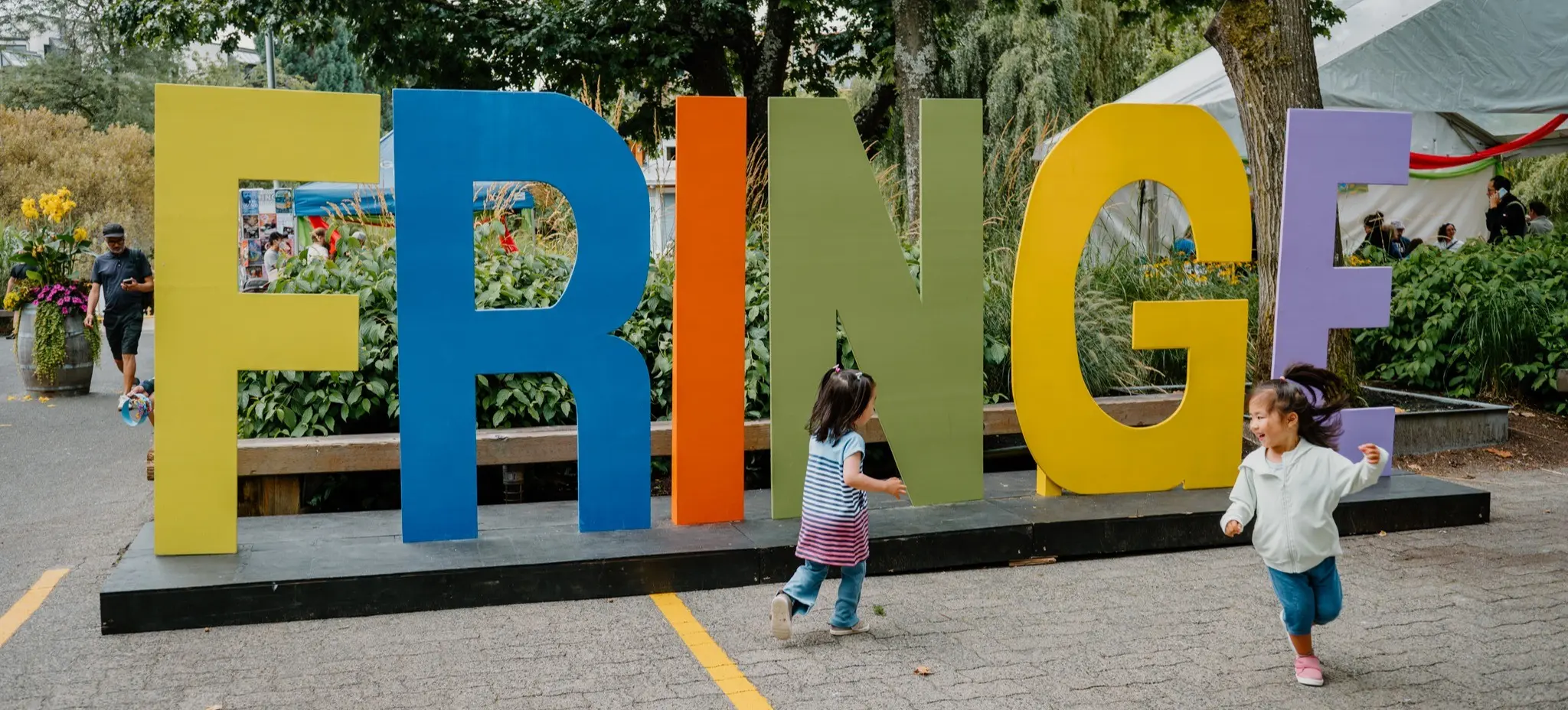 Two children running and playing in front of large colorful letters spelling FRINGE in an outdoor park setting.