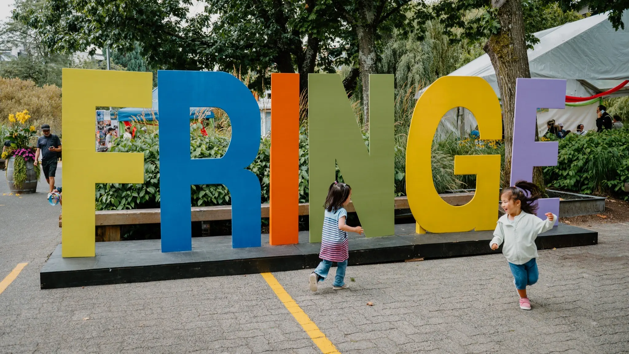 Two children running and playing in front of large colorful letters spelling FRINGE in an outdoor park setting.