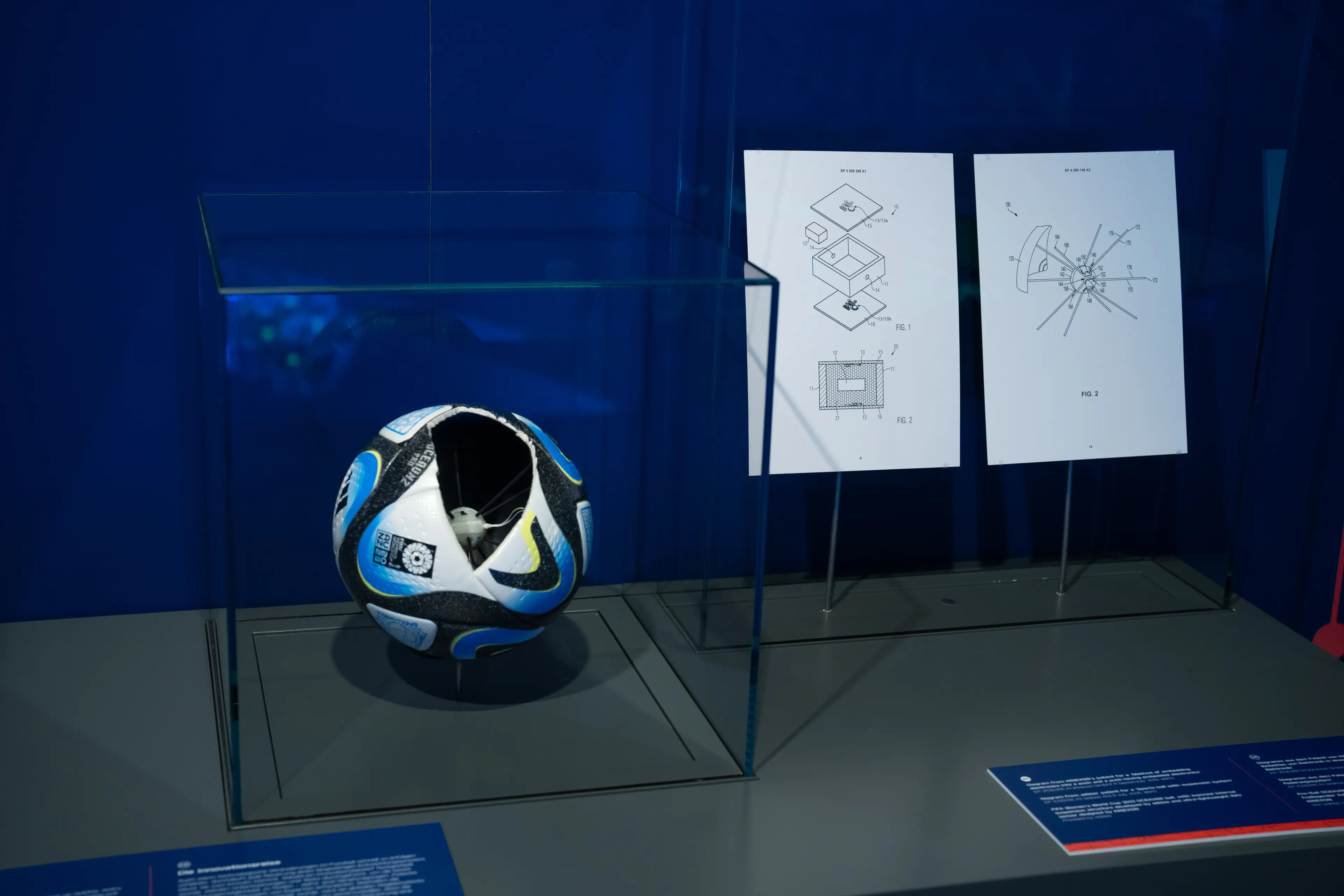 A blue, white, and black soccer ball displayed in a glass case with two technical drawings on white paper behind it.