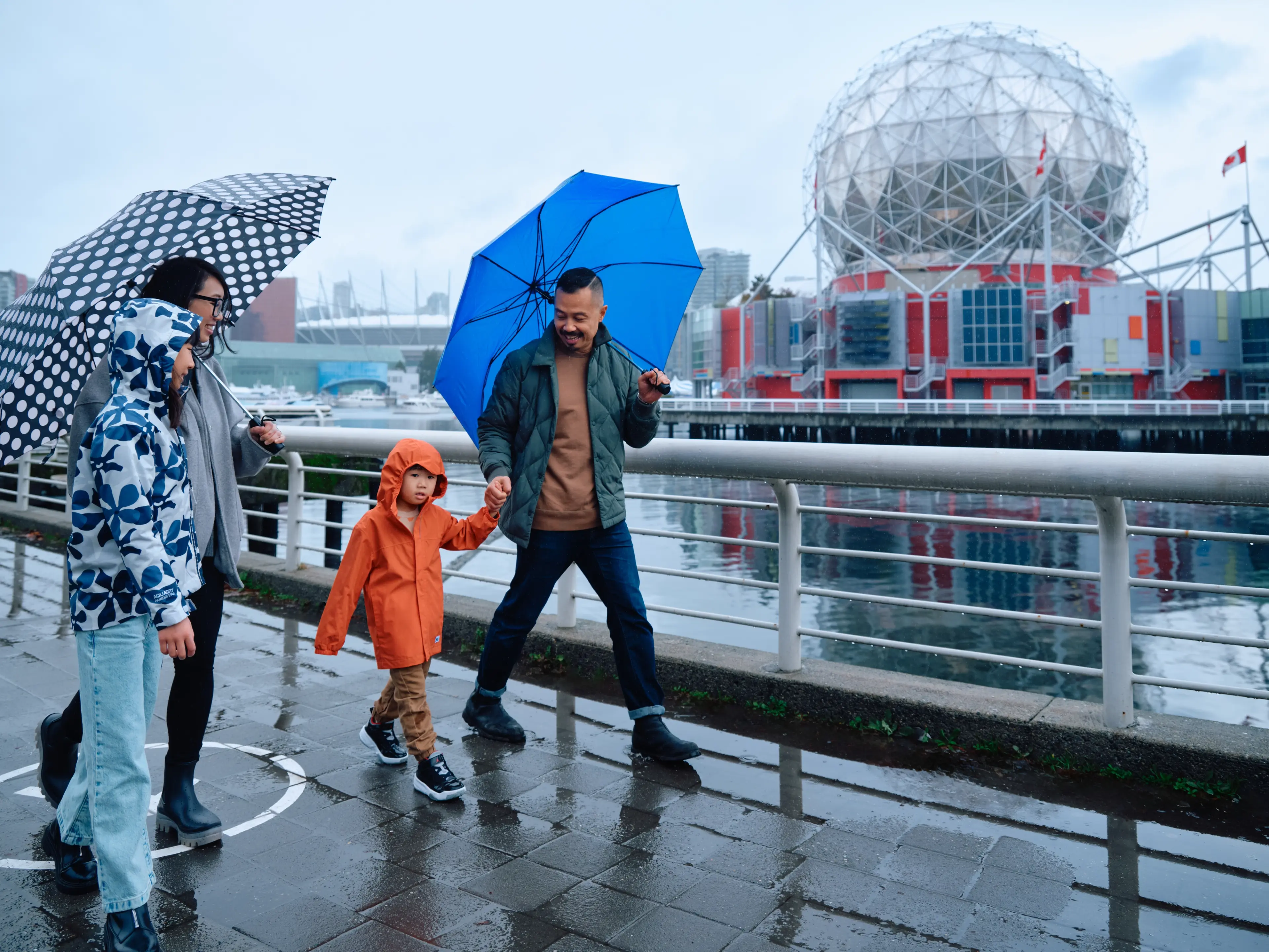 Family walking on a wet waterfront path with umbrellas, a child in an orange raincoat, and a geodesic dome building in the background.