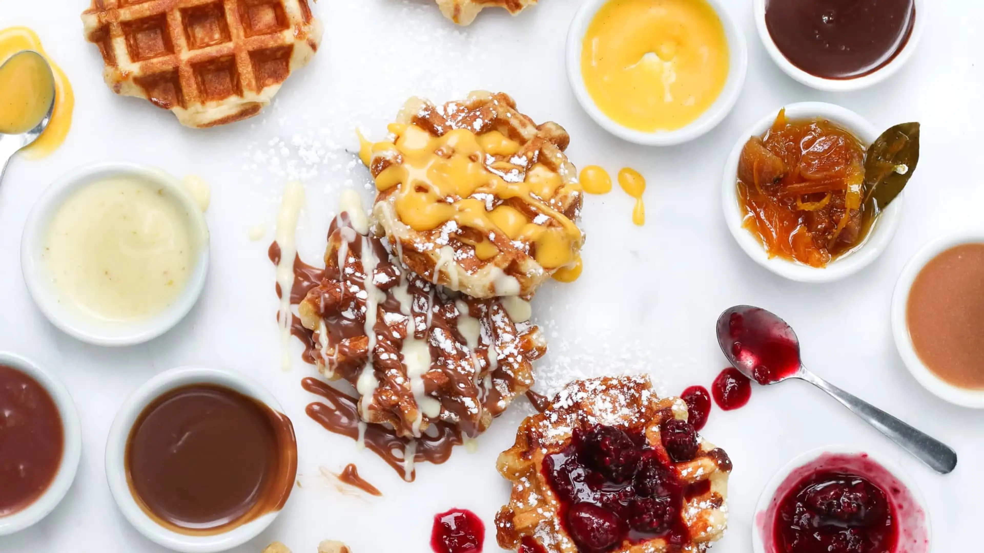 Three waffles topped with yellow sauce, chocolate and white drizzle, and red berry sauce, surrounded by small bowls of various sauces.