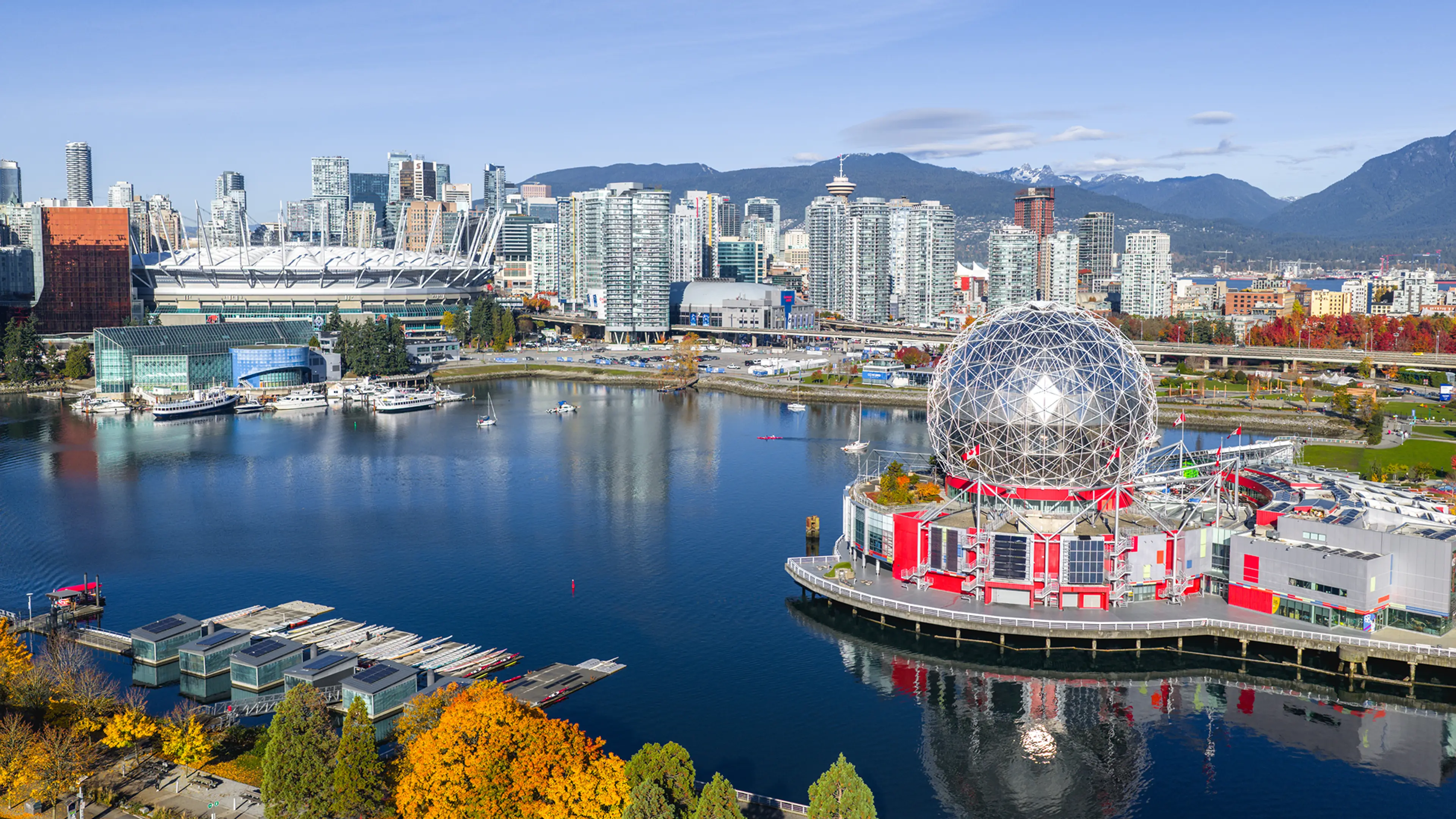 Waterfront cityscape with a geodesic dome building, boats on water, and mountains in the background.