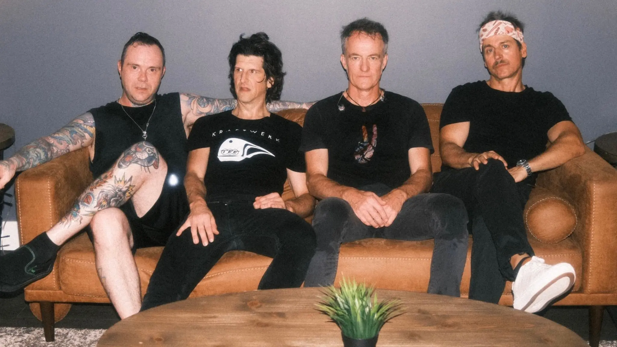 Four people sitting on a brown leather couch against a gray wall, with a wooden table and a small green plant in front.