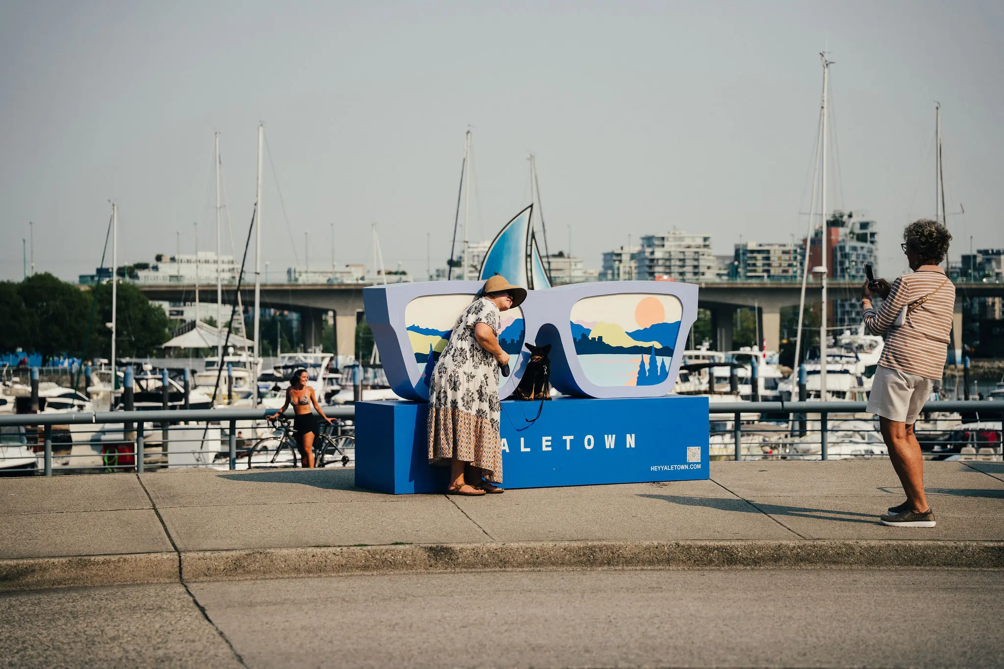 Woman in a hat posing with a dog inside a large blue sunglasses sculpture at a marina while another person takes a photo.