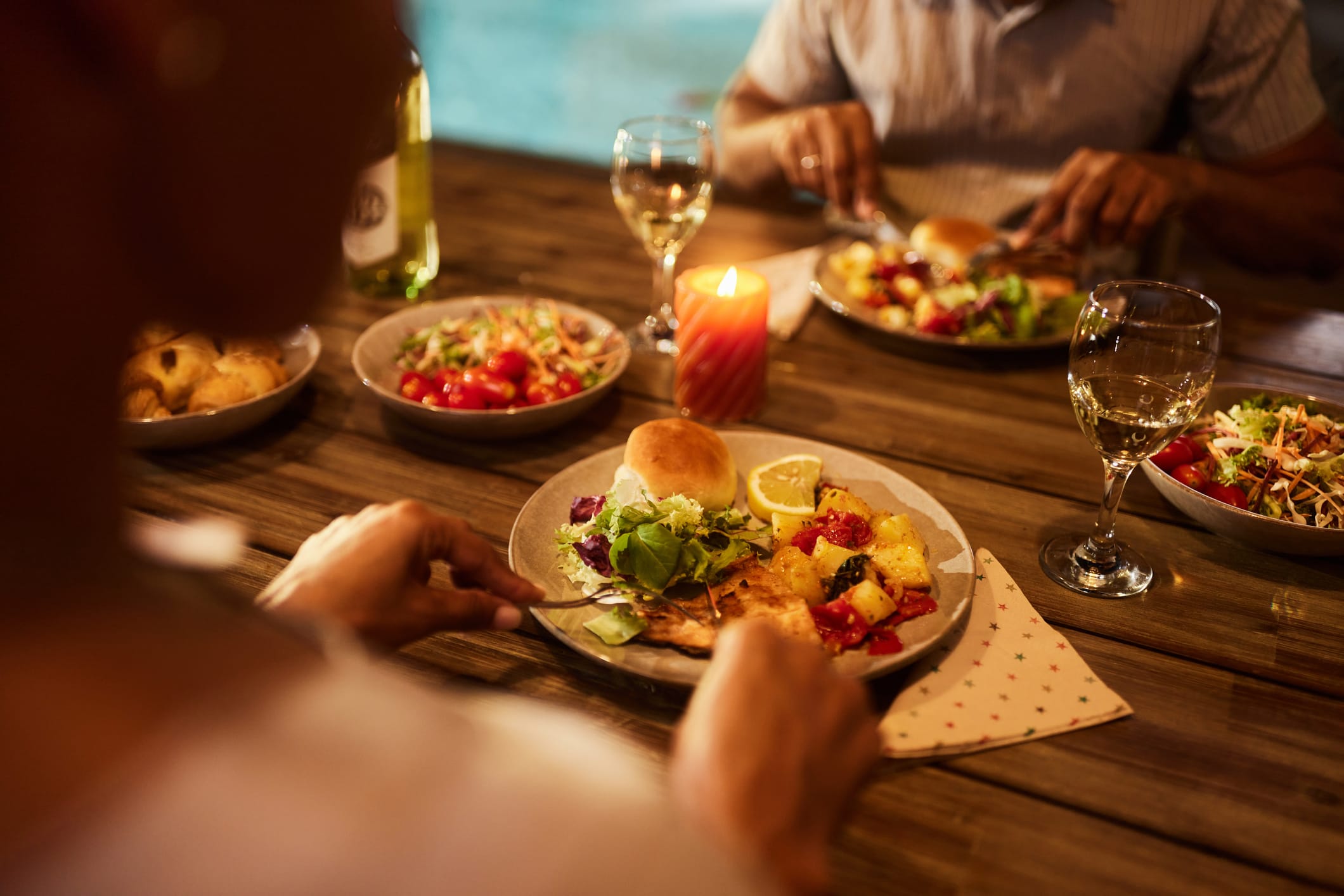 Two people dining at a wooden table with plates of salad, bread roll, grilled food, and glasses of white wine, lit by a candle.