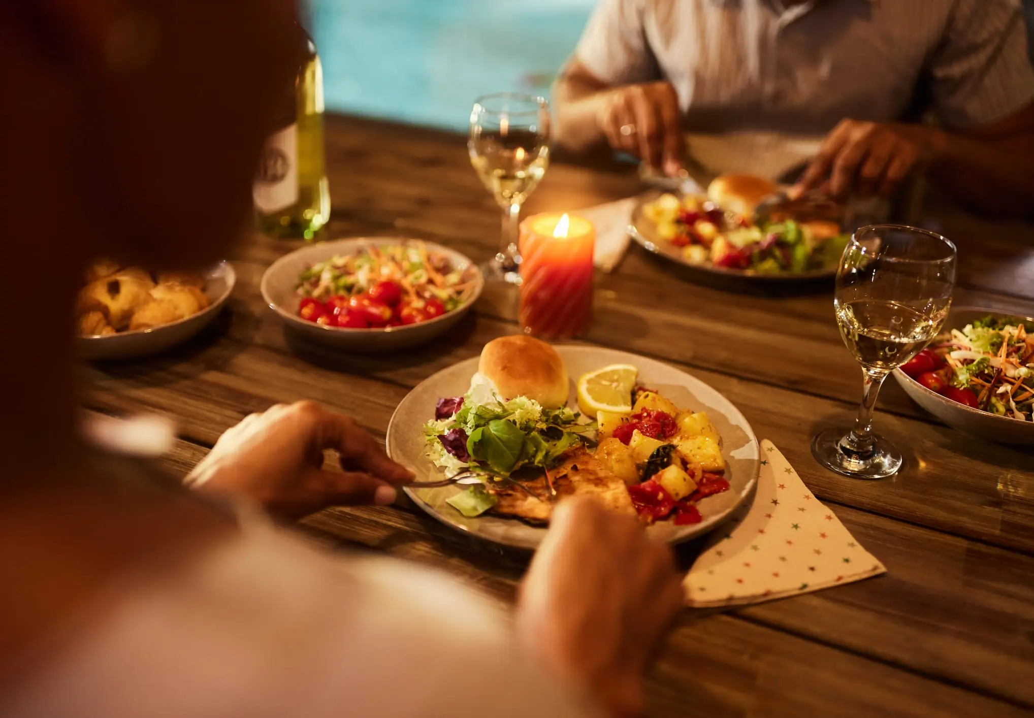 Two people dining at a wooden table with plates of salad, bread roll, grilled food, and glasses of white wine, lit by a candle.