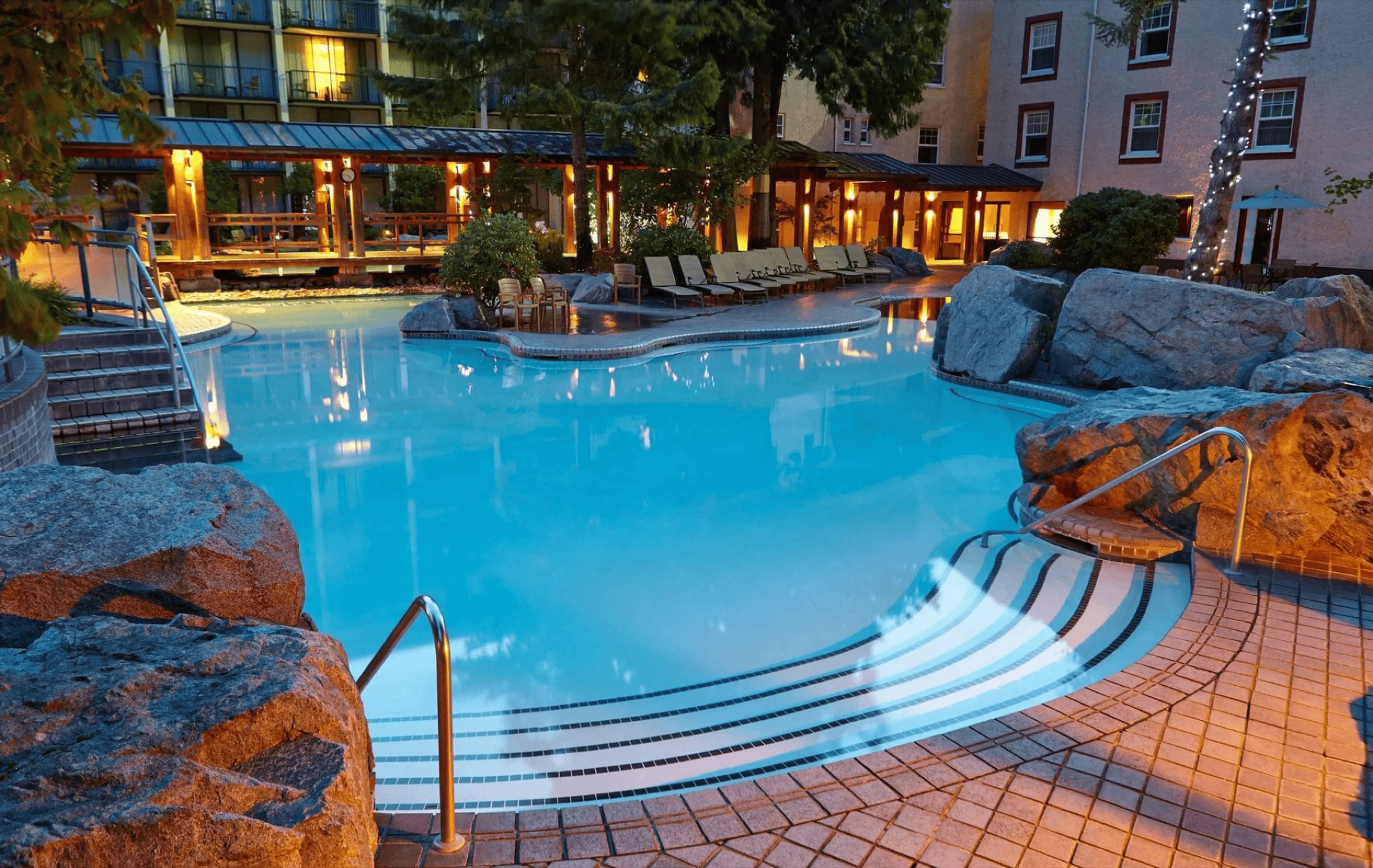 A serene outdoor swimming pool at dusk, surrounded by rocks, lounge chairs, and warm lighting from nearby buildings—an ideal retreat for those seeking relaxing day trips from Vancouver in a tranquil, inviting atmosphere.