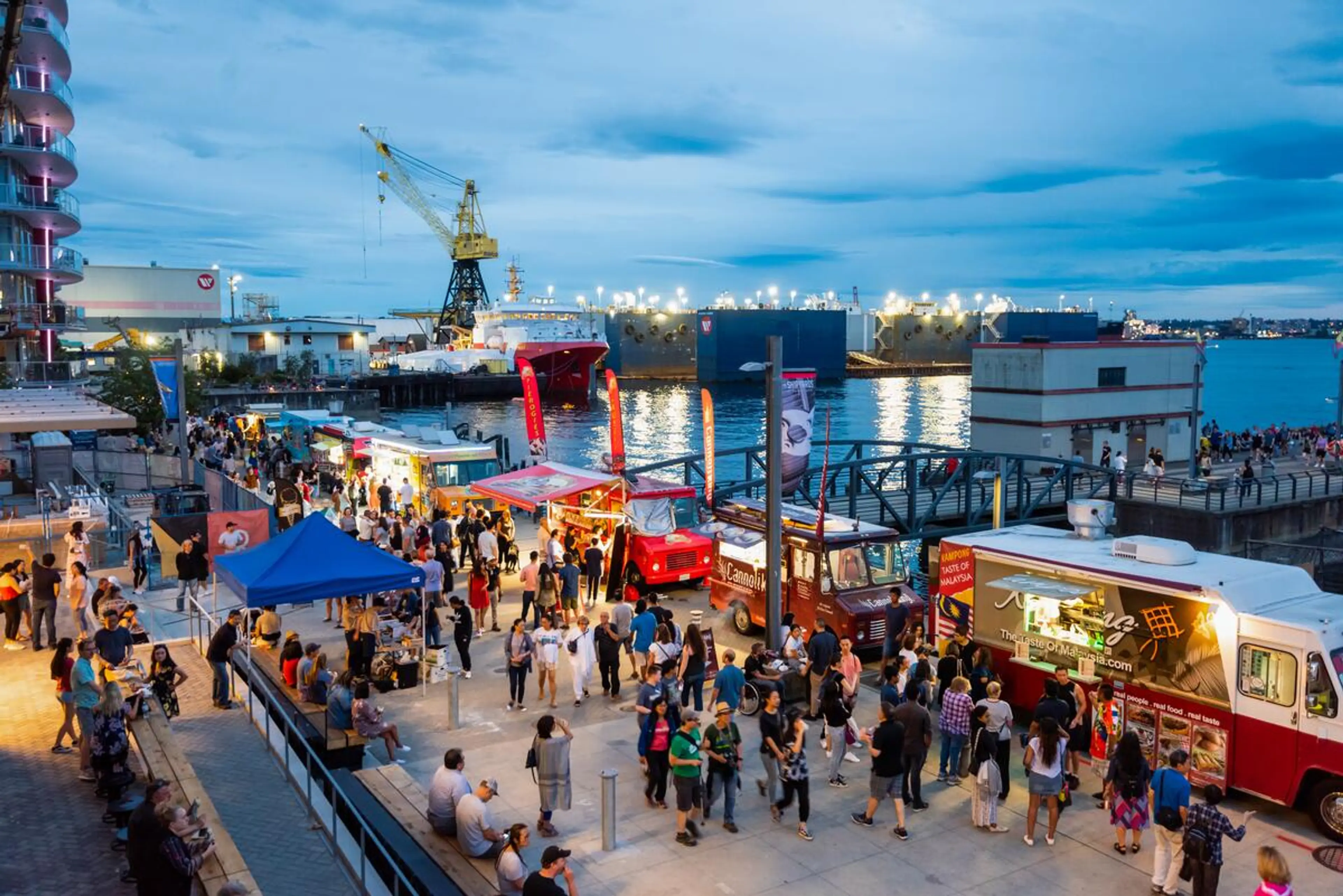 Crowded waterfront food truck festival at dusk with people walking and sitting near the water and boats in the background at the Shipyards Night Market, Vancouver.