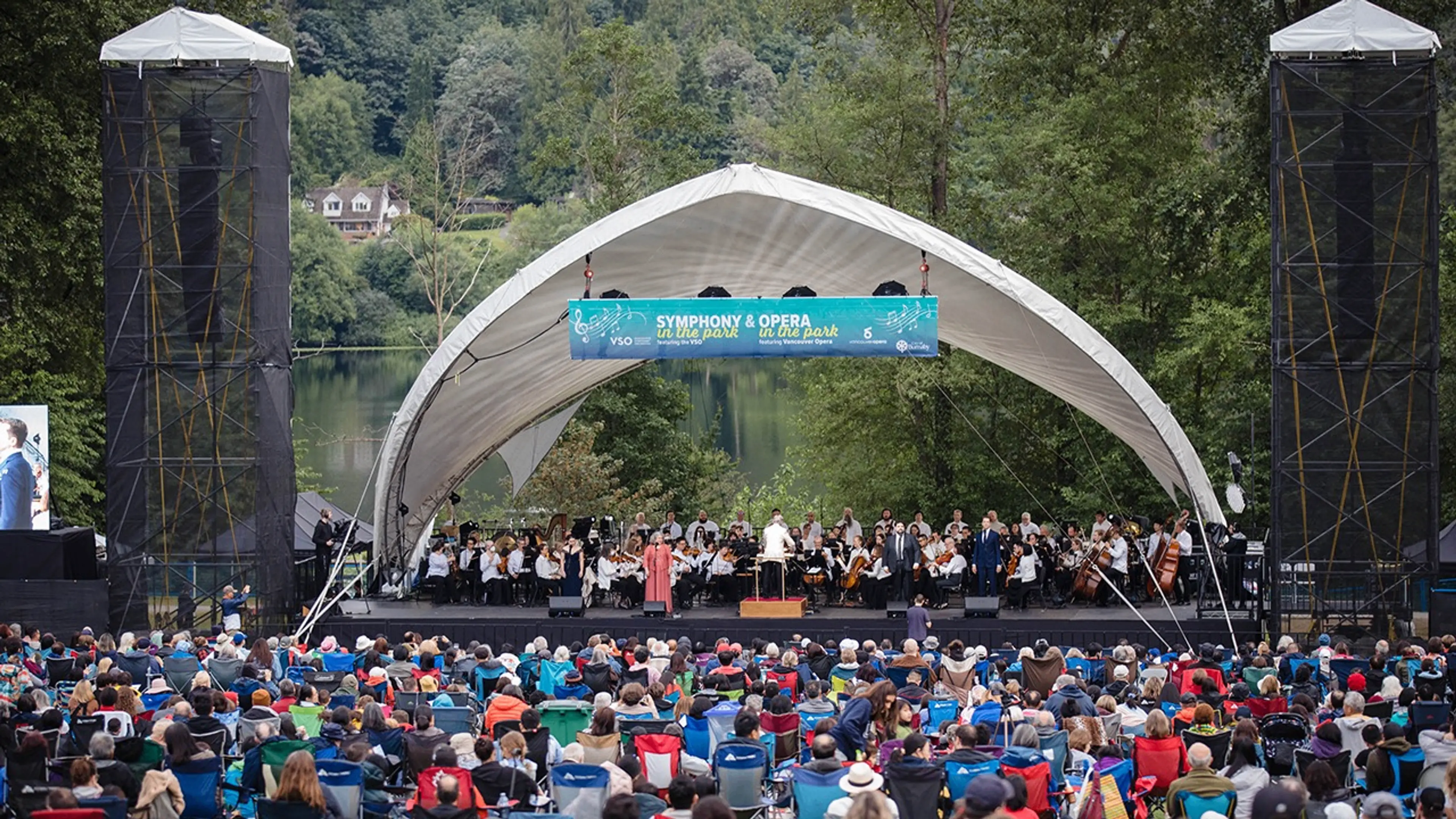 Outdoor symphony and opera concert with orchestra and singers on stage, audience seated on lawn chairs in park.