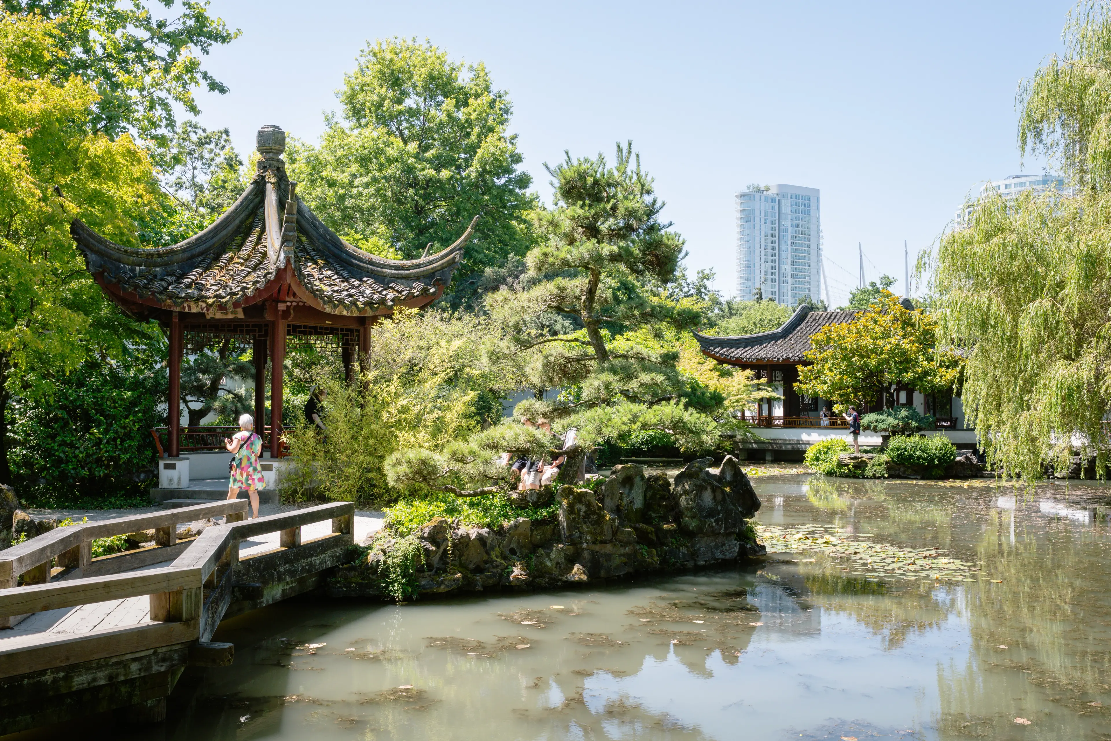 Traditional Chinese garden with a pond, wooden bridge, pagoda, and people walking among lush green trees.